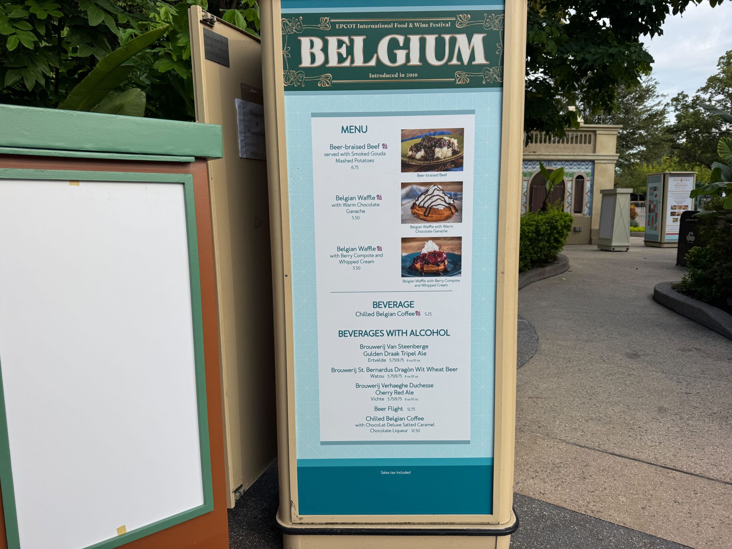 Sign for the Belgium booth displays Food & Wine menu items and beverages at an outdoor festival, with trees in the background.