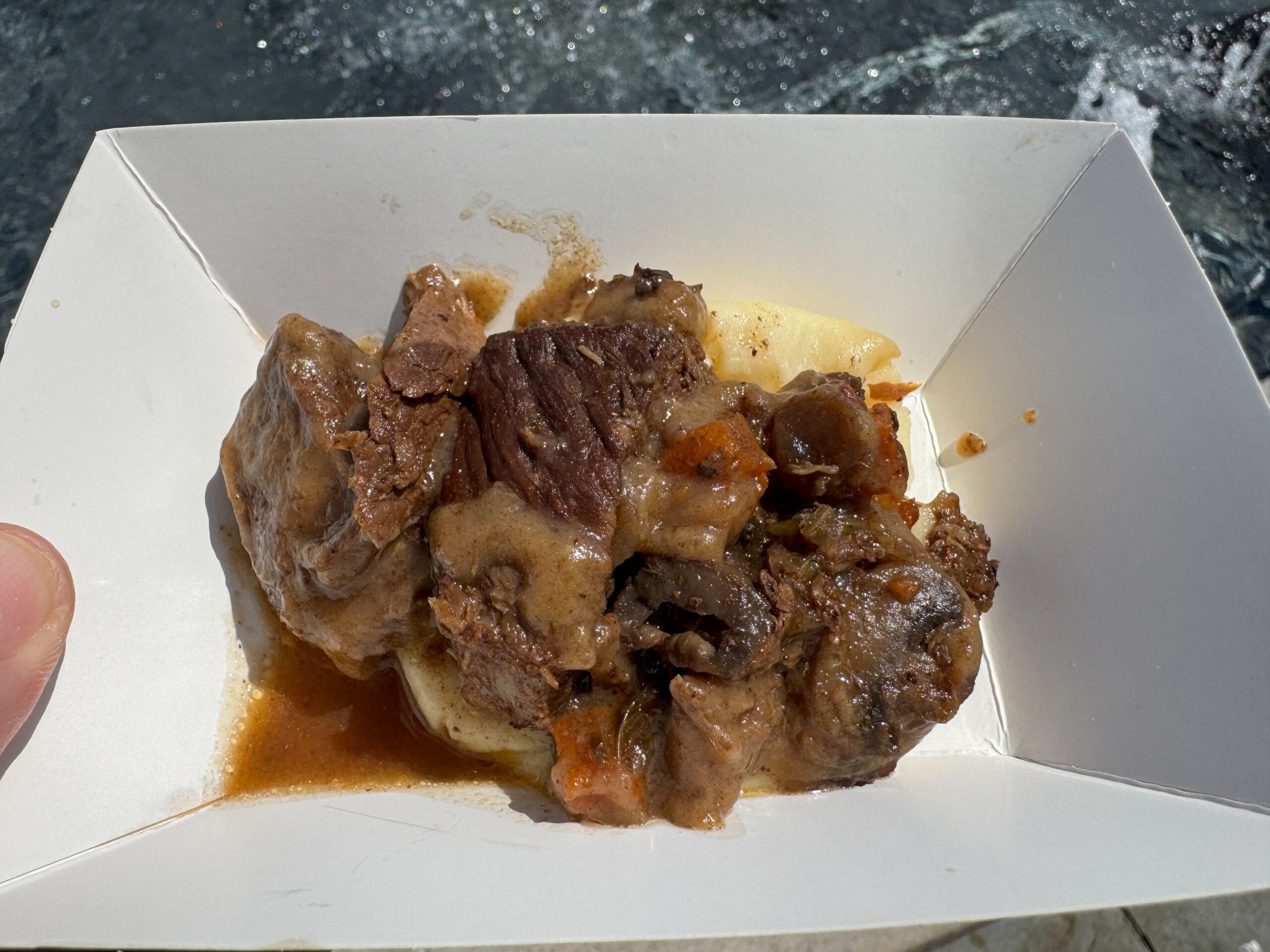 Beef stew with sauce and vegetables served over mashed potatoes in a white paper tray—a Food & Wine favorite.