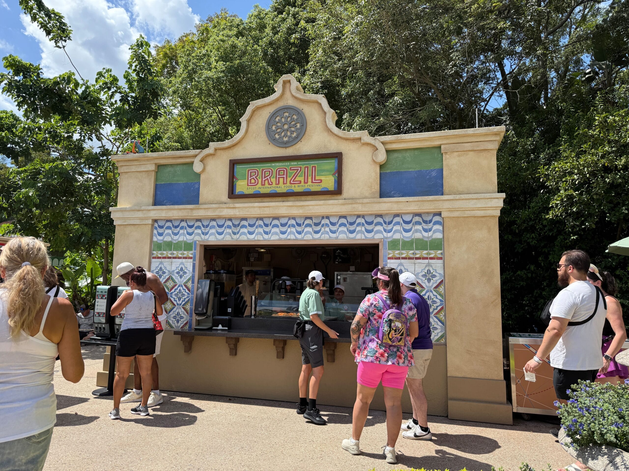People wait in line at an outdoor Food & Wine stand labeled "Brazil," surrounded by trees and colorful tiles.