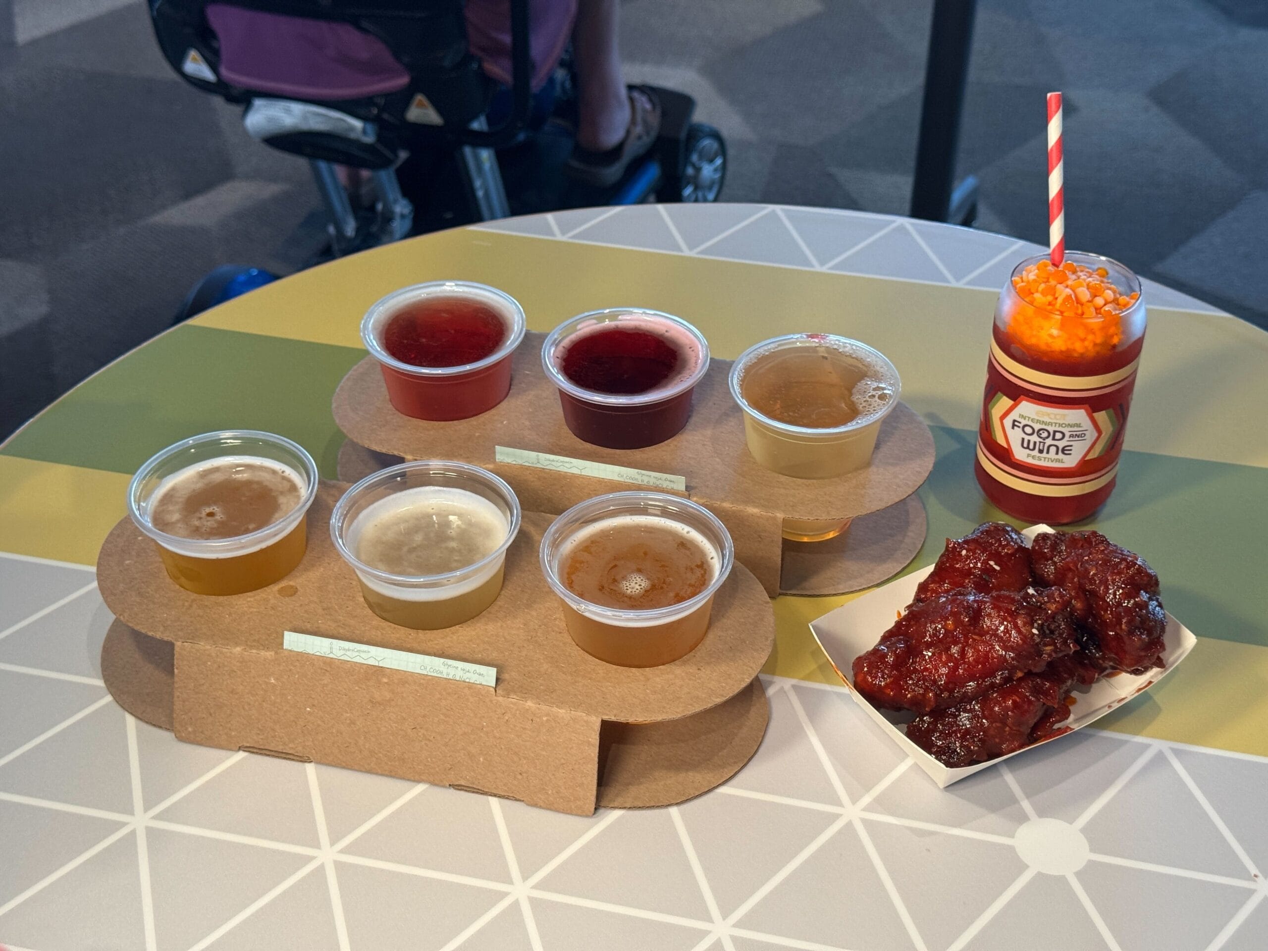 A Food & Wine flight of six beers, sticky glazed wings, and a themed orange drink on a geometric table.
