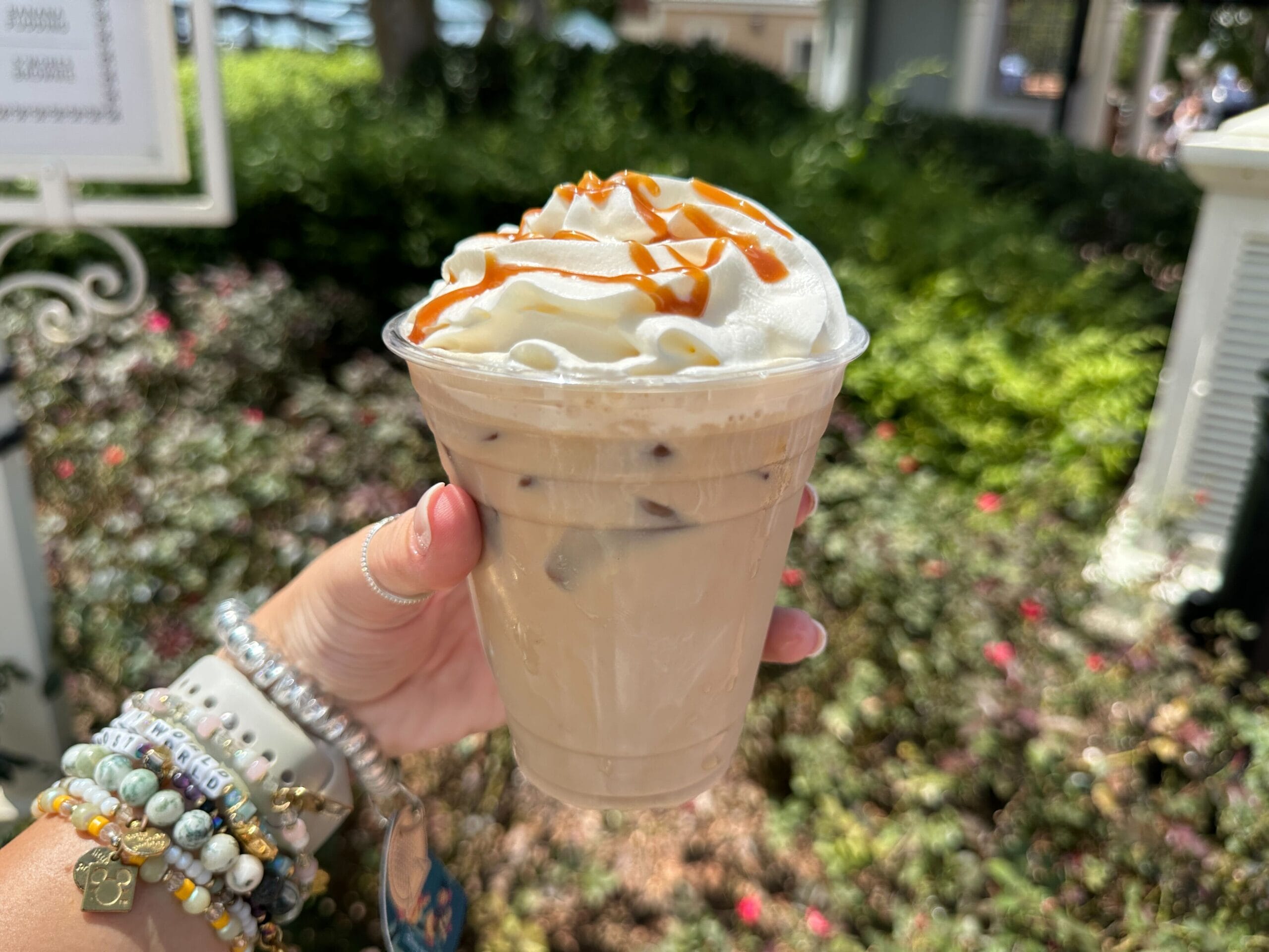 Hand holding an iced coffee topped with whipped cream and caramel drizzle, perfect for Food & Wine lovers in a garden setting.
