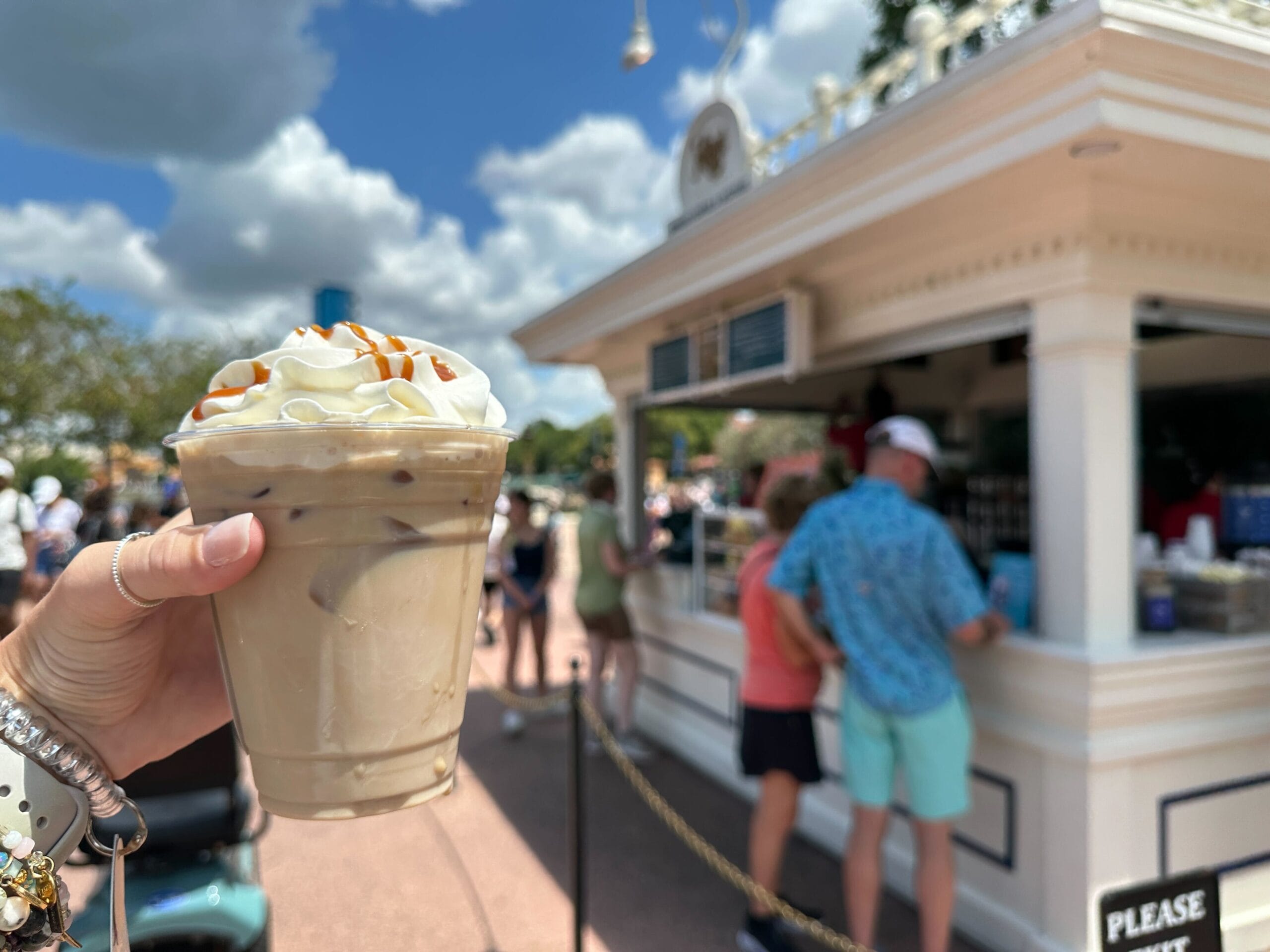 A hand holds an iced coffee with whipped cream and caramel, Food & Wine style, in front of an outdoor café on a sunny day.