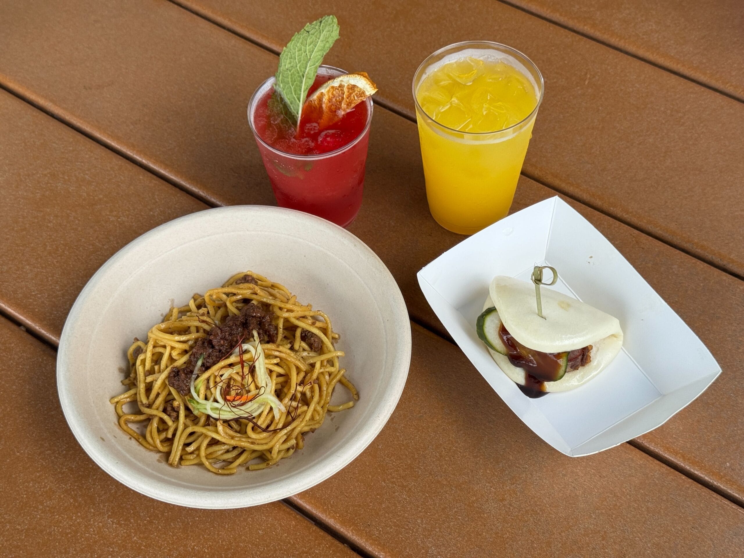 A bowl of noodles, a bao bun, and colorful drinks on a wooden table at the EPCOT Food and Wine Festival.