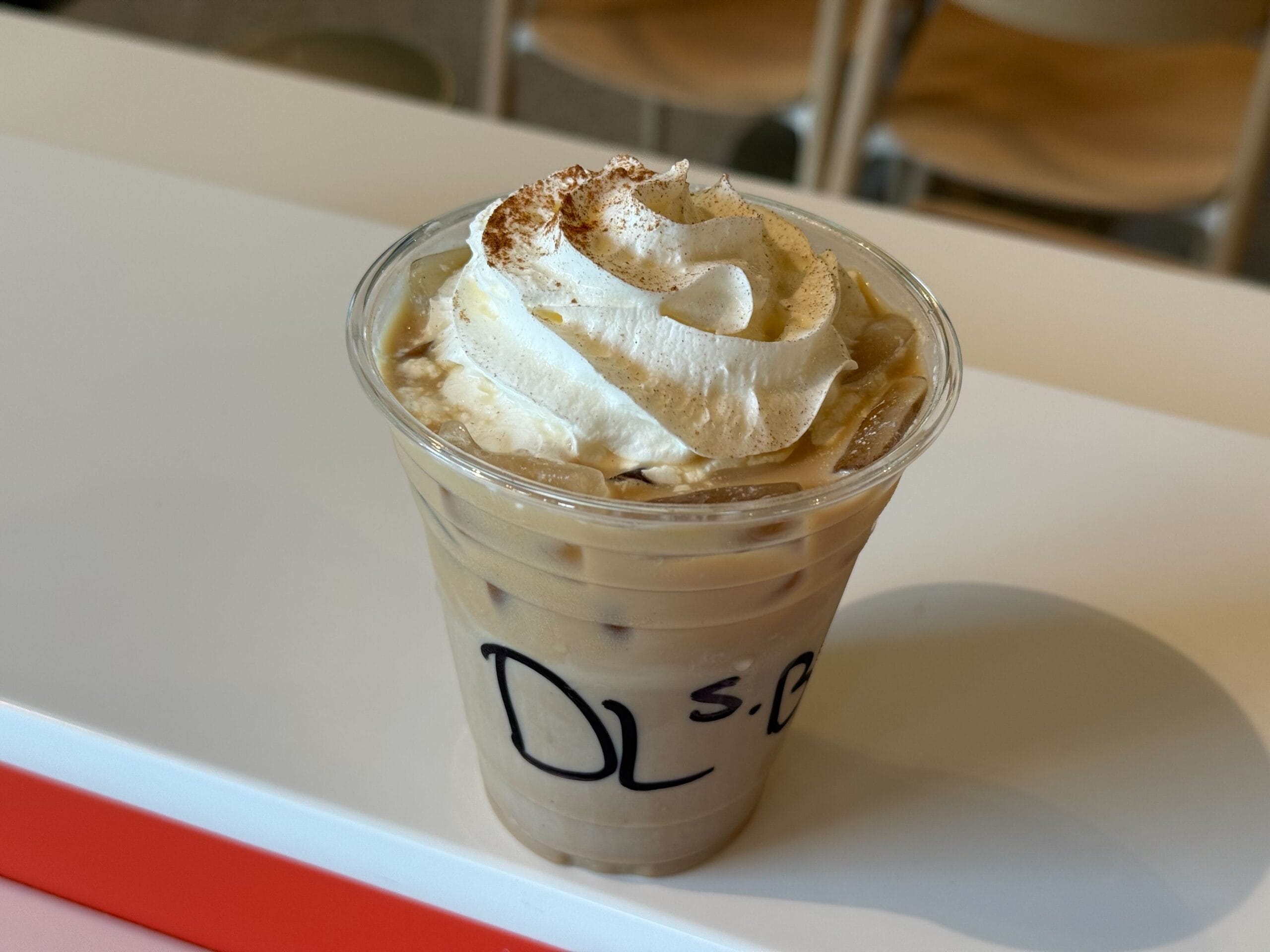 Iced coffee in a clear plastic cup with whipped cream and cinnamon, a refreshing New Food & Wine treat on a white table.