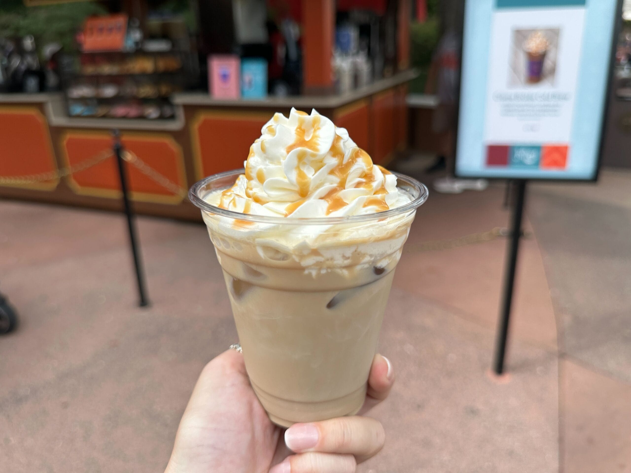 A hand holds an iced coffee topped with whipped cream and caramel, perfect for a Food & Wine break at an outdoor café.