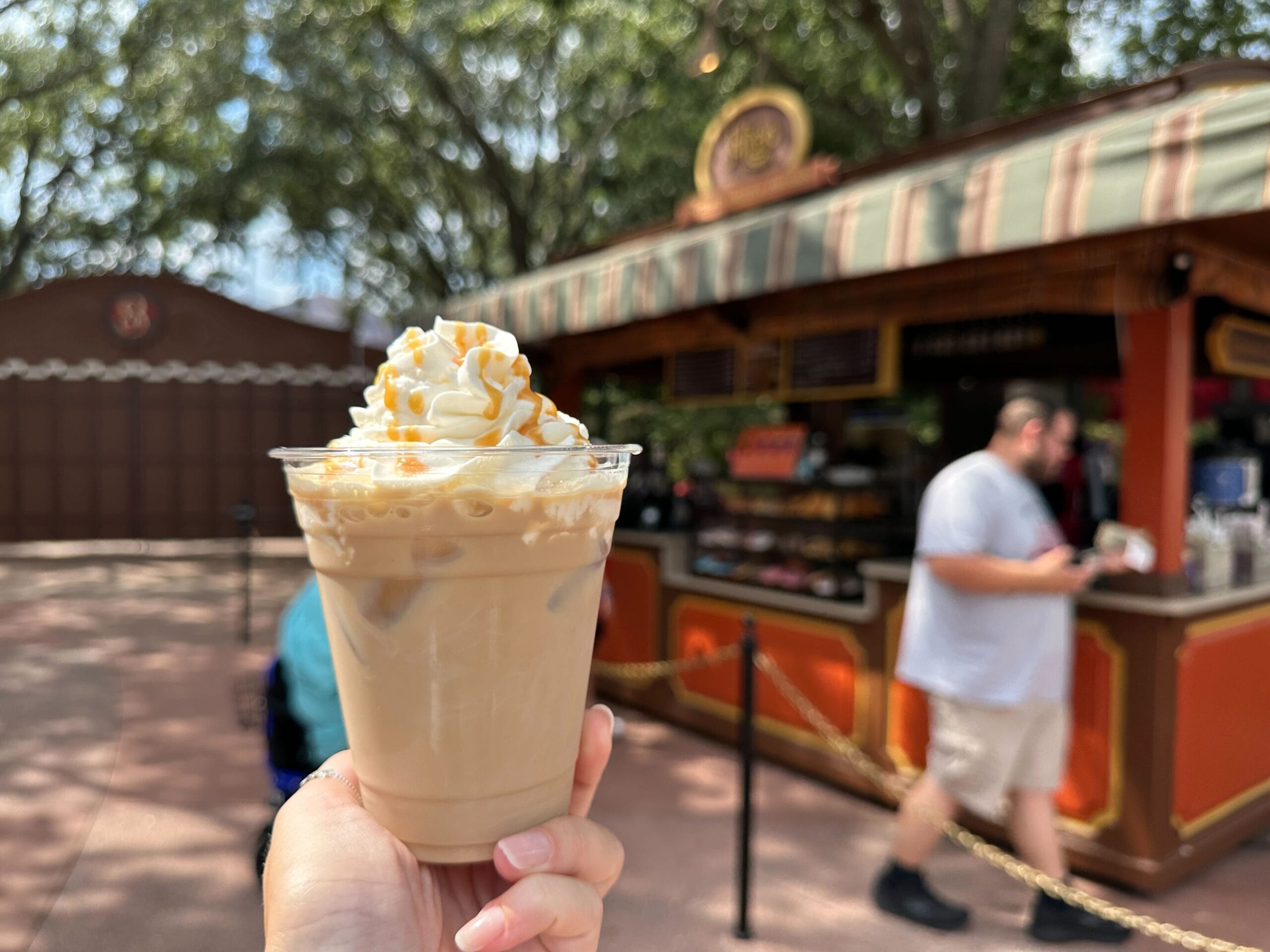 A hand holds an iced coffee topped with whipped cream and caramel at a vibrant Food & Wine outdoor stand.
