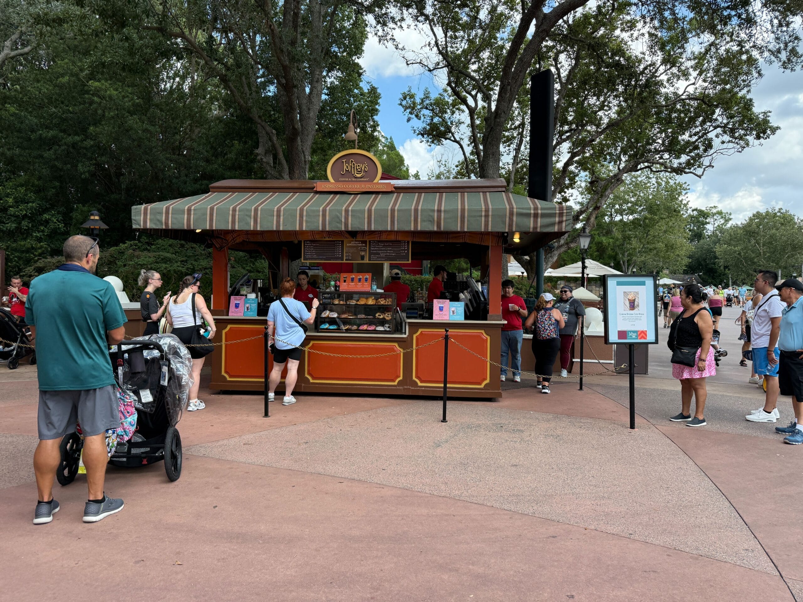 People stand in line at a New Food & Wine kiosk with a striped canopy and menu signs at a park.
