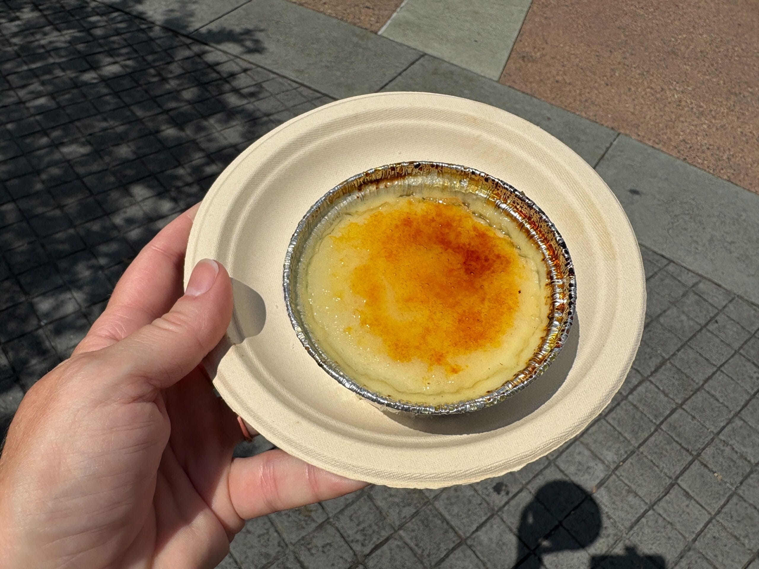 A hand holds a plate with crème brûlée in a foil dish—Food & Wine enjoyed outdoors on a sunny day.