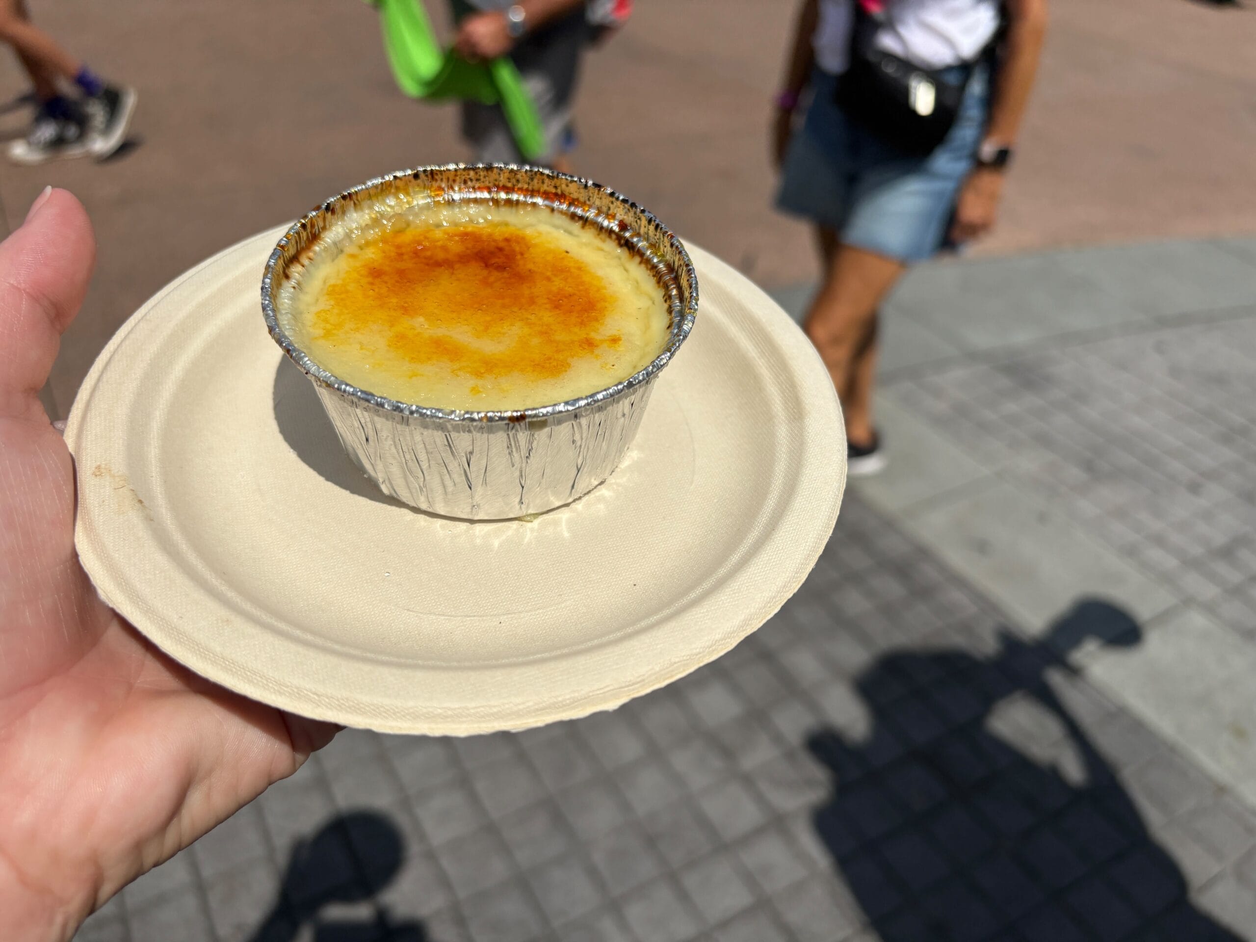 A hand holds a plate with crème brûlée in a foil cup at a Food & Wine event; people and shadows appear behind.