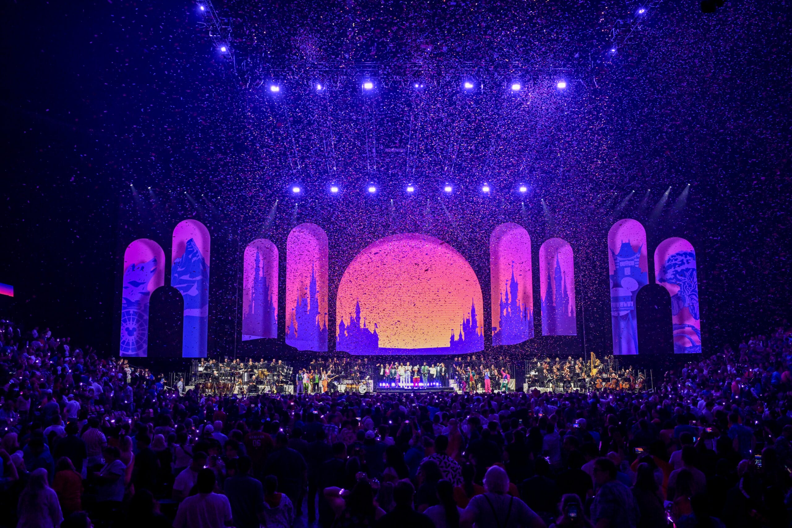 A vibrant concert stage at the 2026 D23 Disney Fan Event, with a sunset backdrop, arches, confetti, and a lively crowd.