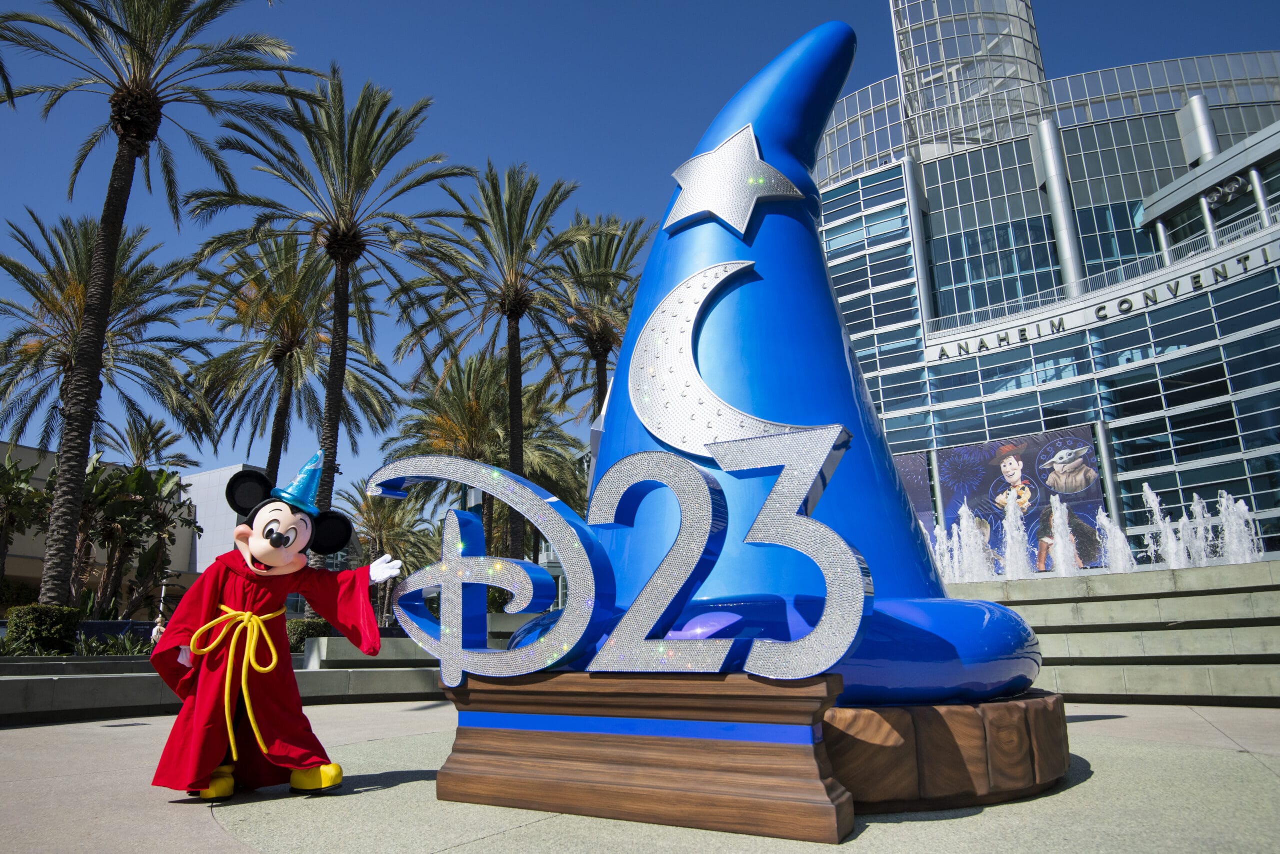 Mickey Mouse in a red robe stands by a large "D23" sign, celebrating the Disney Fan Event outside Anaheim Convention Center.