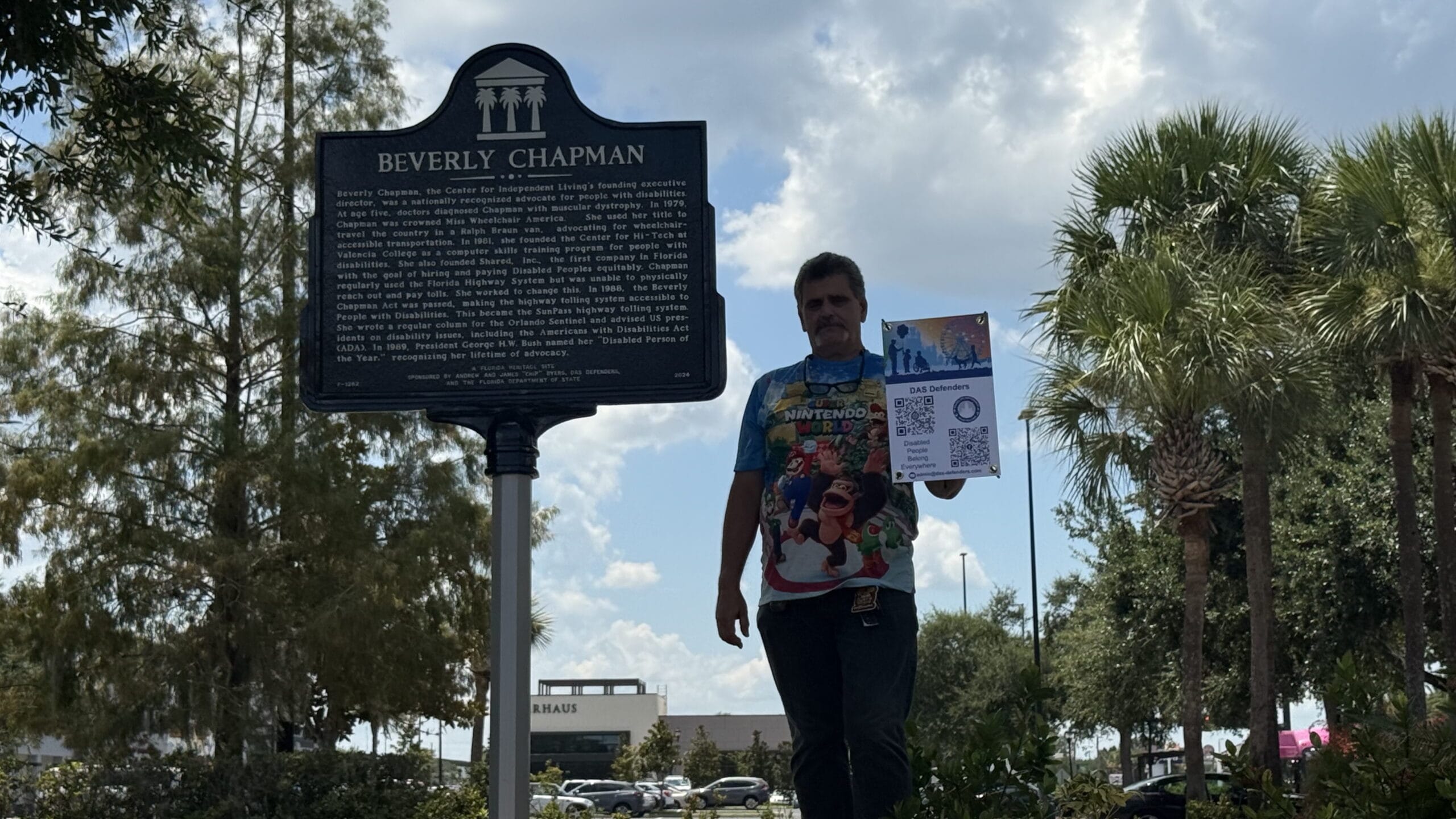 A person stands by the Beverly Chapman historical marker, holding a sign, with trees and a building in the background.