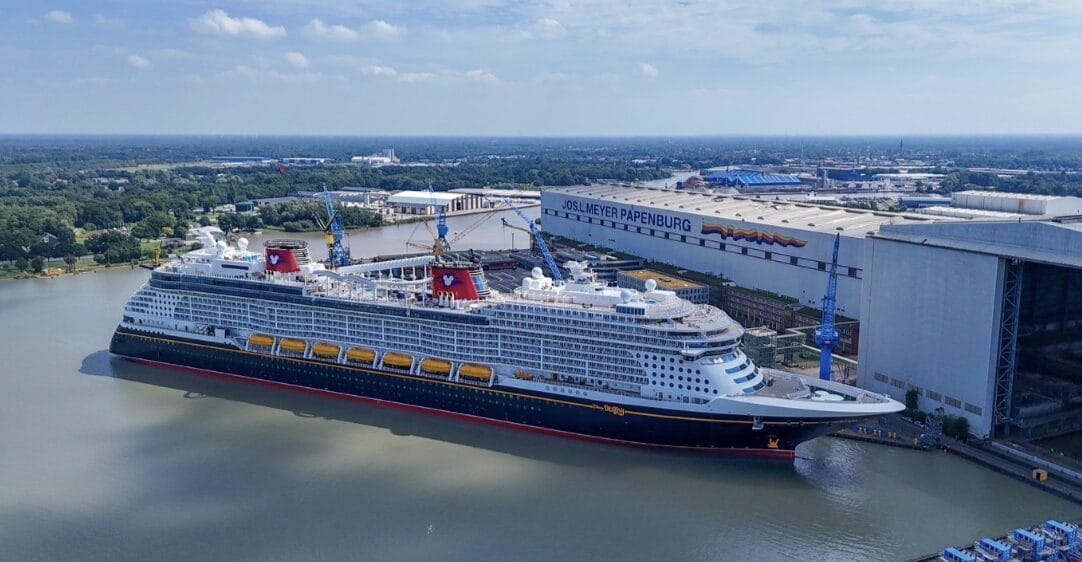 Large cruise ship docked beside the Meyer Werft shipyard in Papenburg, Germany, on a sunny day.