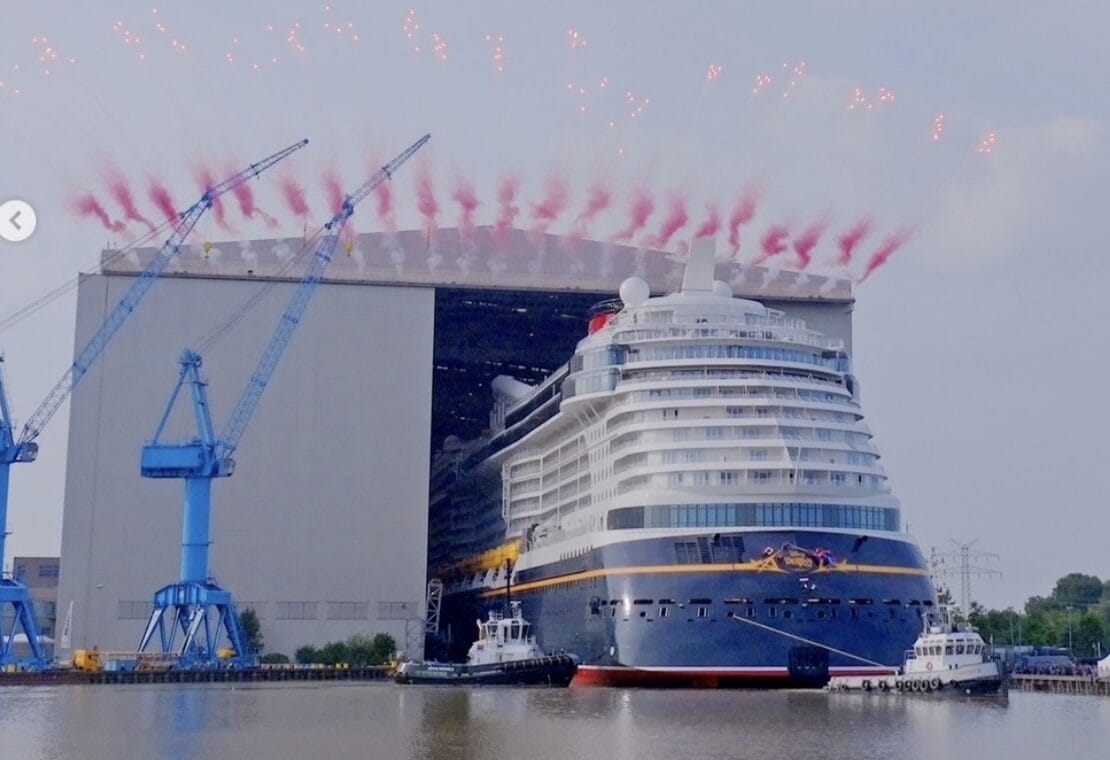 A large cruise ship leaves a shipyard as red fireworks burst above and tugboats guide it on the water.