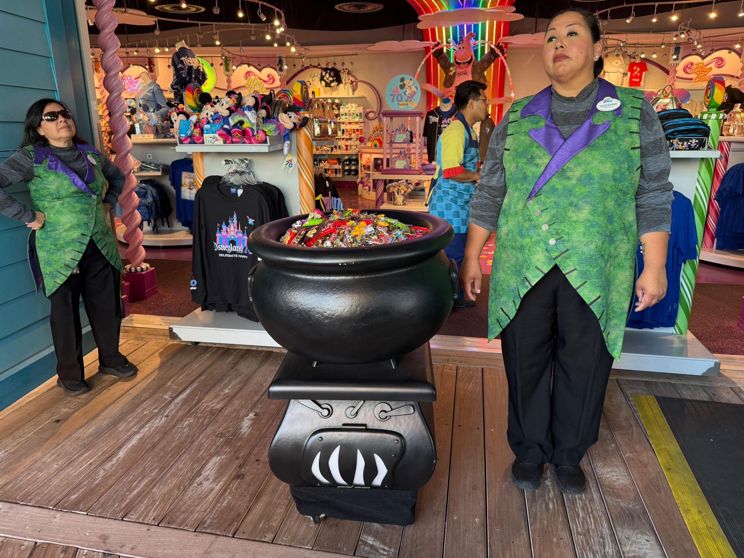 Two employees in green vests stand by a cauldron of candy at a Disney Treat Trail Location inside a colorful store.