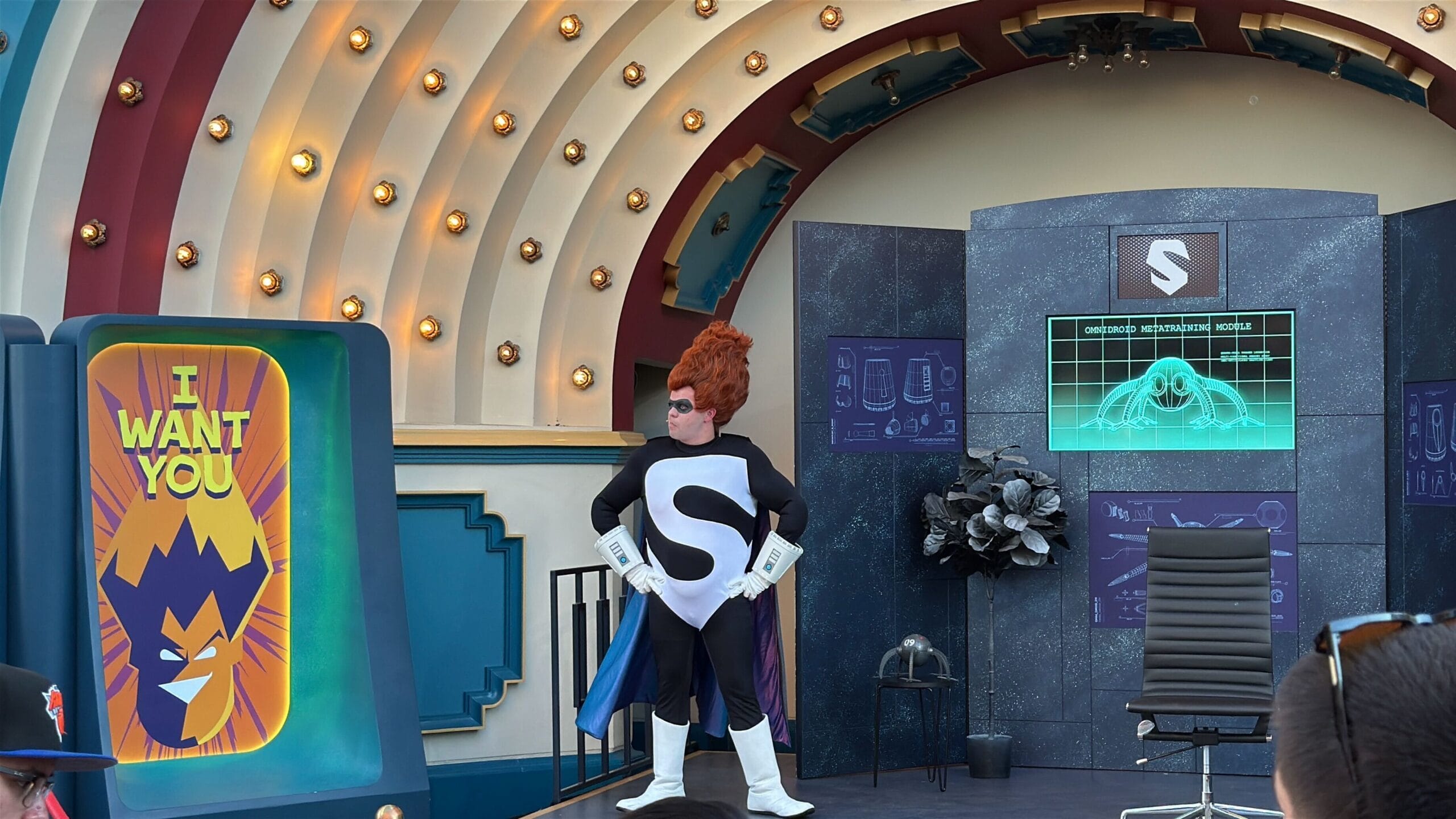 A person in a superhero costume stands on stage at Oogie Boogie Bash beside a villain's "I WANT YOU" poster.