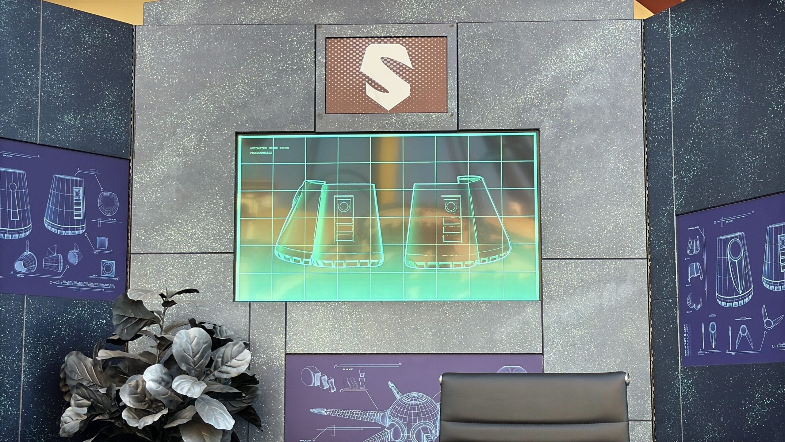 A futuristic screen displays blueprints on a gray wall at Disney, with plant decor and a black chair in front.