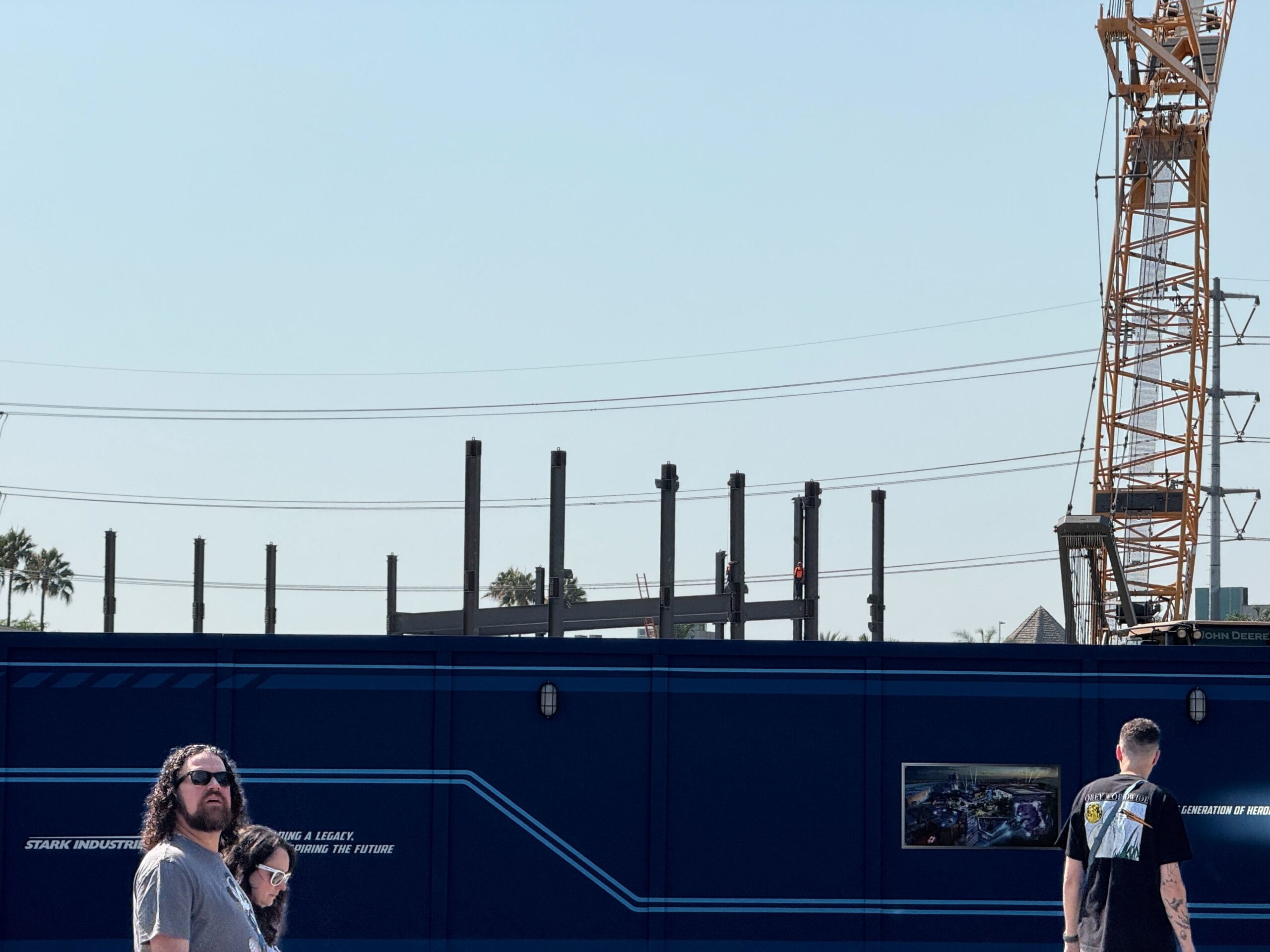 People walk in front of a blue construction fence in Avengers Campus; steel beams and a crane are visible behind the fence under a clear sky.