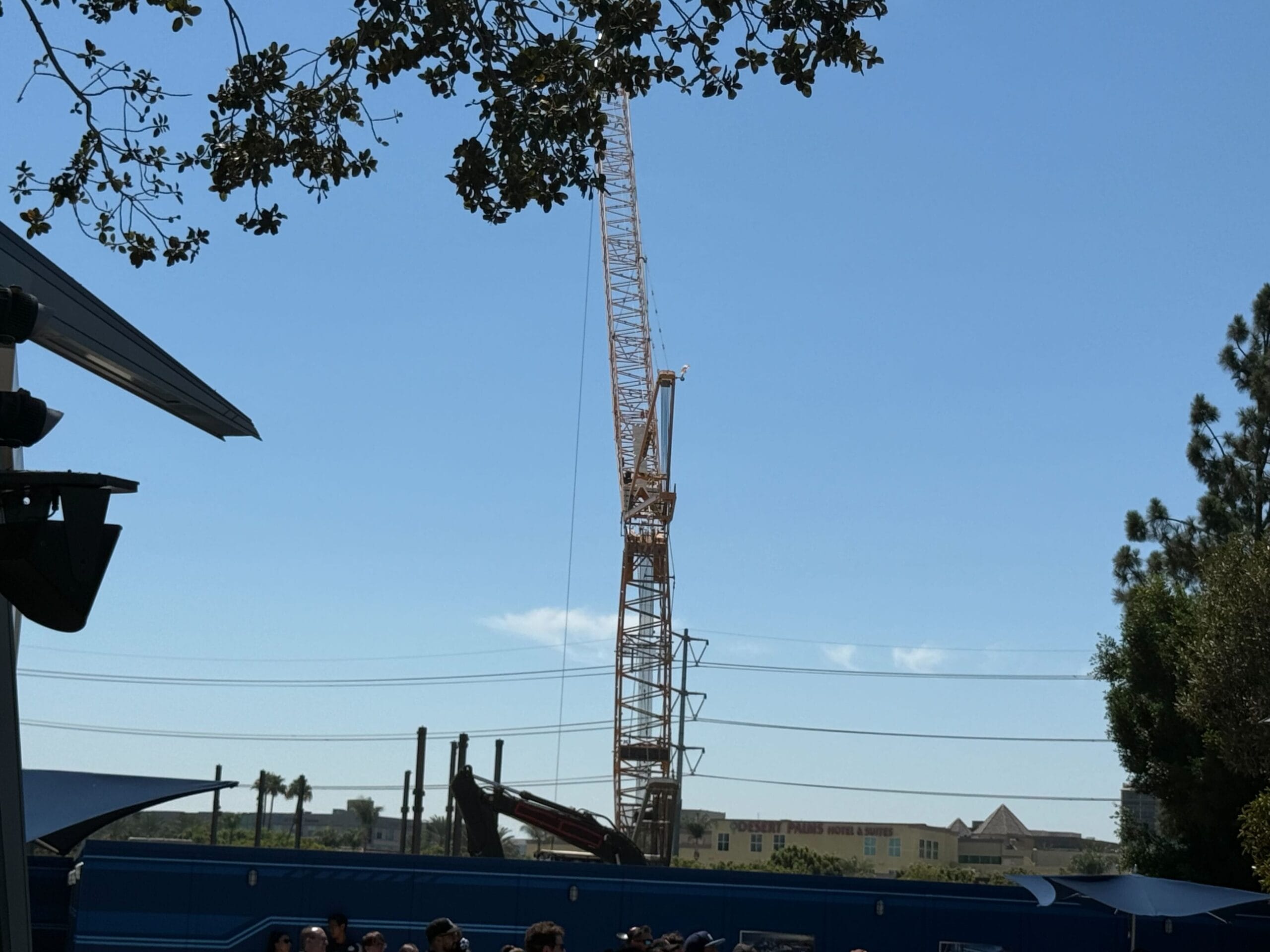 A tall construction crane stands in Avengers Campus against a blue sky, with trees and a crowd in the foreground.