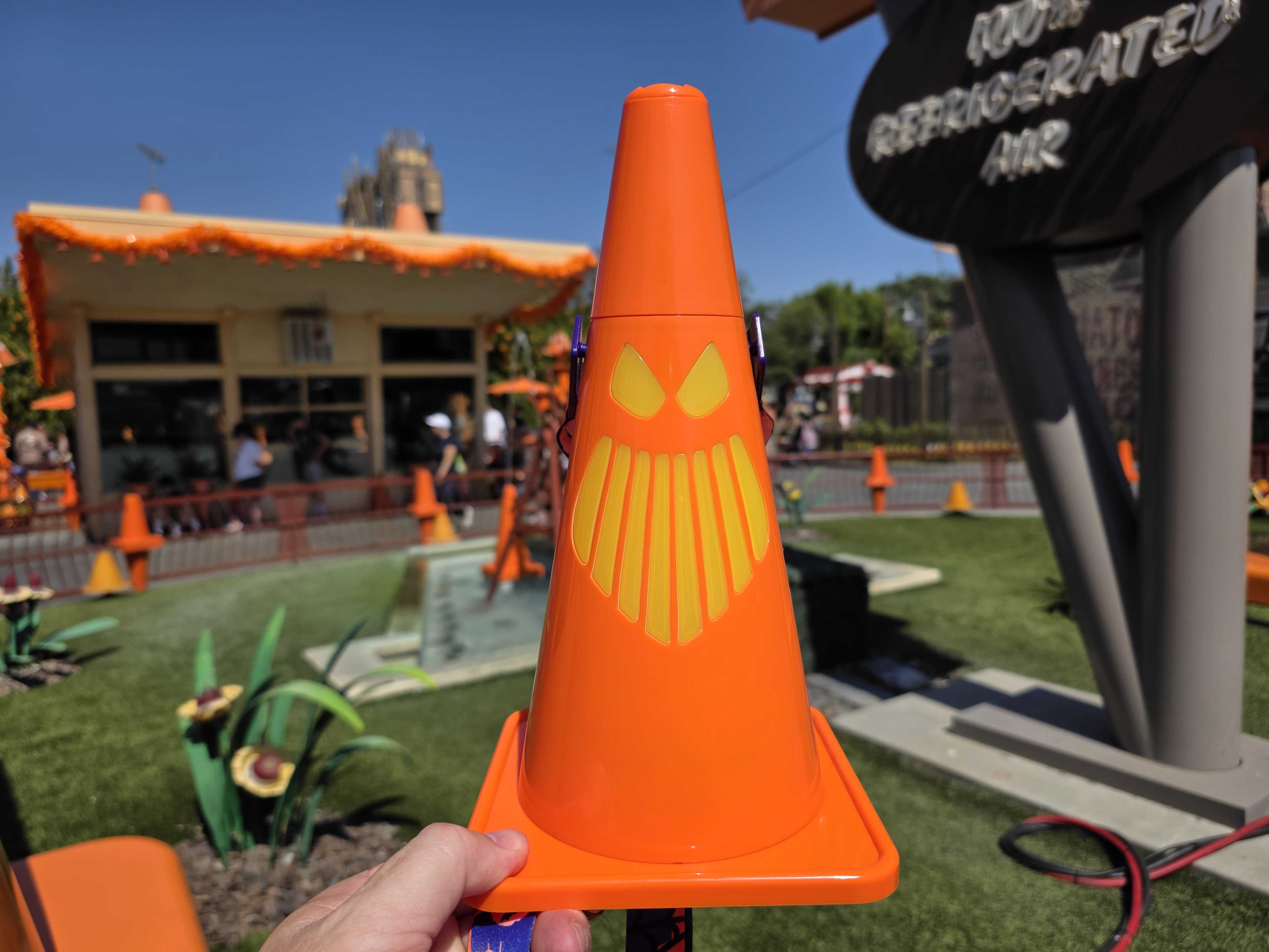 An orange traffic cone with a yellow, sinister face design is held up in an outdoor, theme park setting.