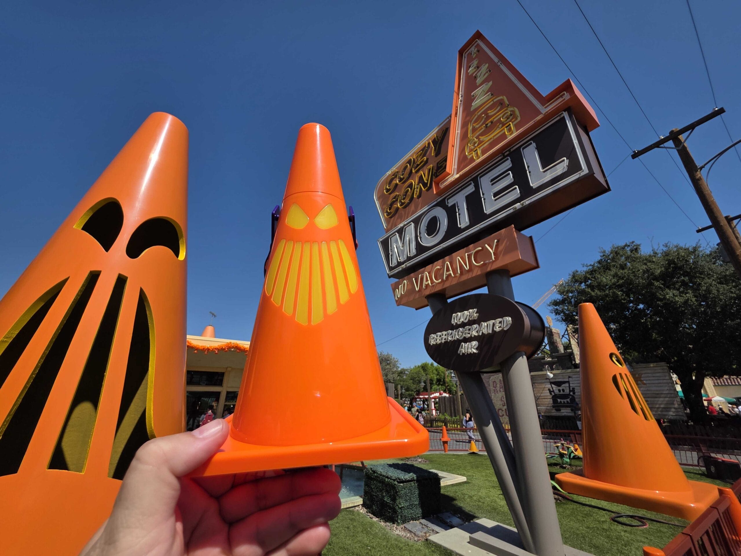 A hand holds an orange cone with a face near a retro motel sign and large, decorated cones outside.