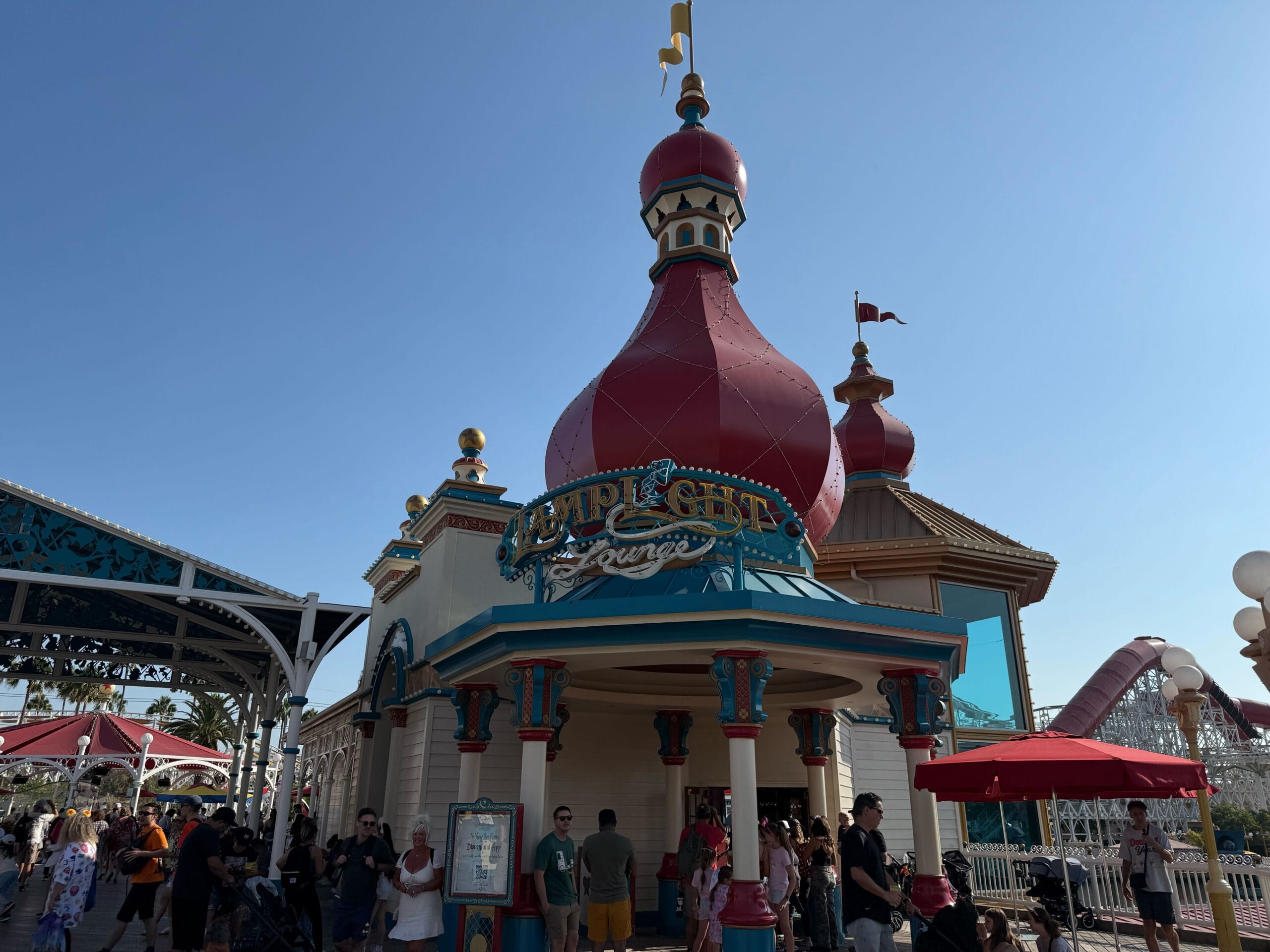 People gather outside the whimsical Lamplight Lounge with its red domes at a theme park on a sunny day.