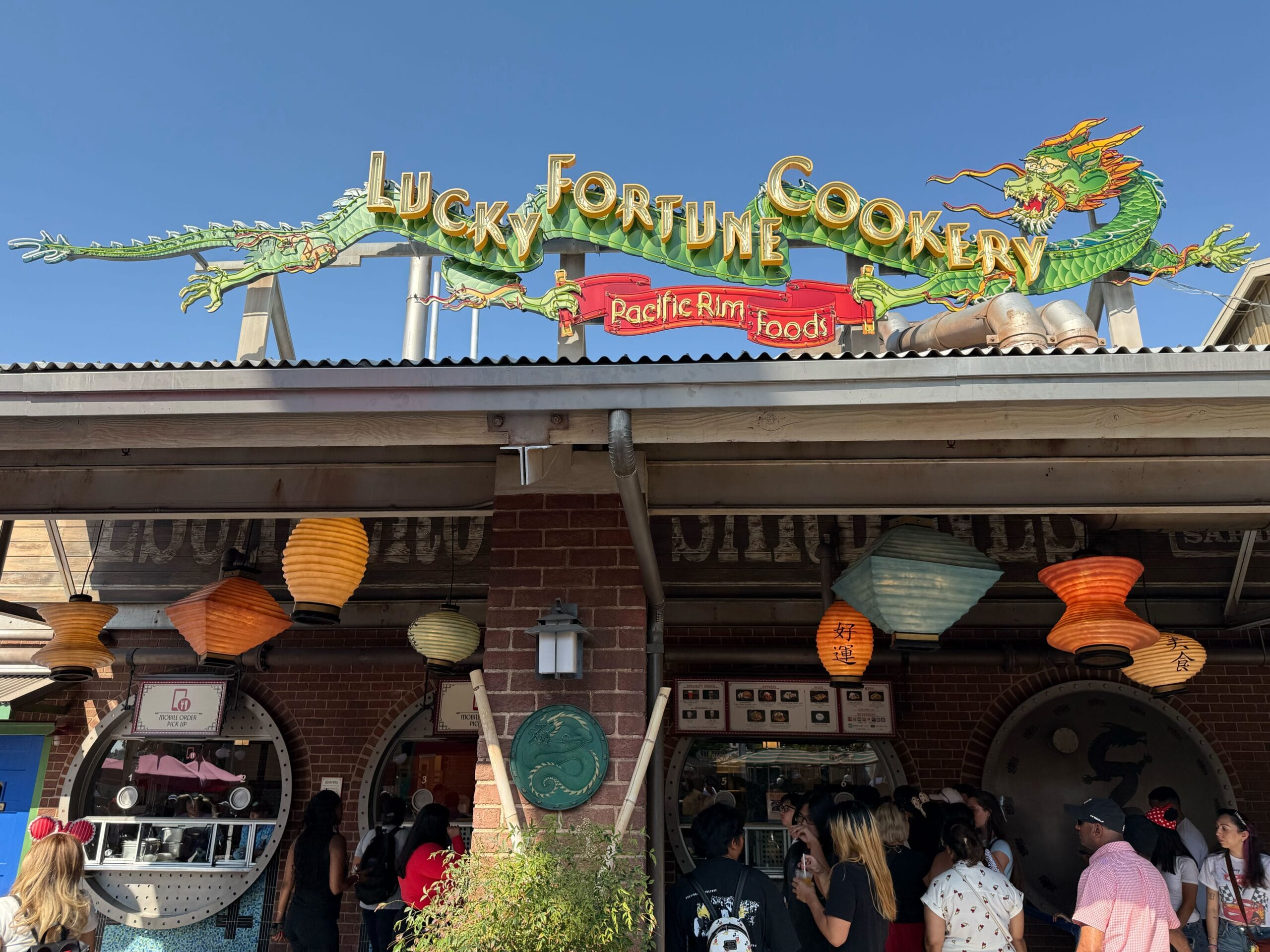 Crowd stands outside Lucky Fortune Cookery restaurant, decorated with a dragon sign and colorful lanterns under a blue sky.