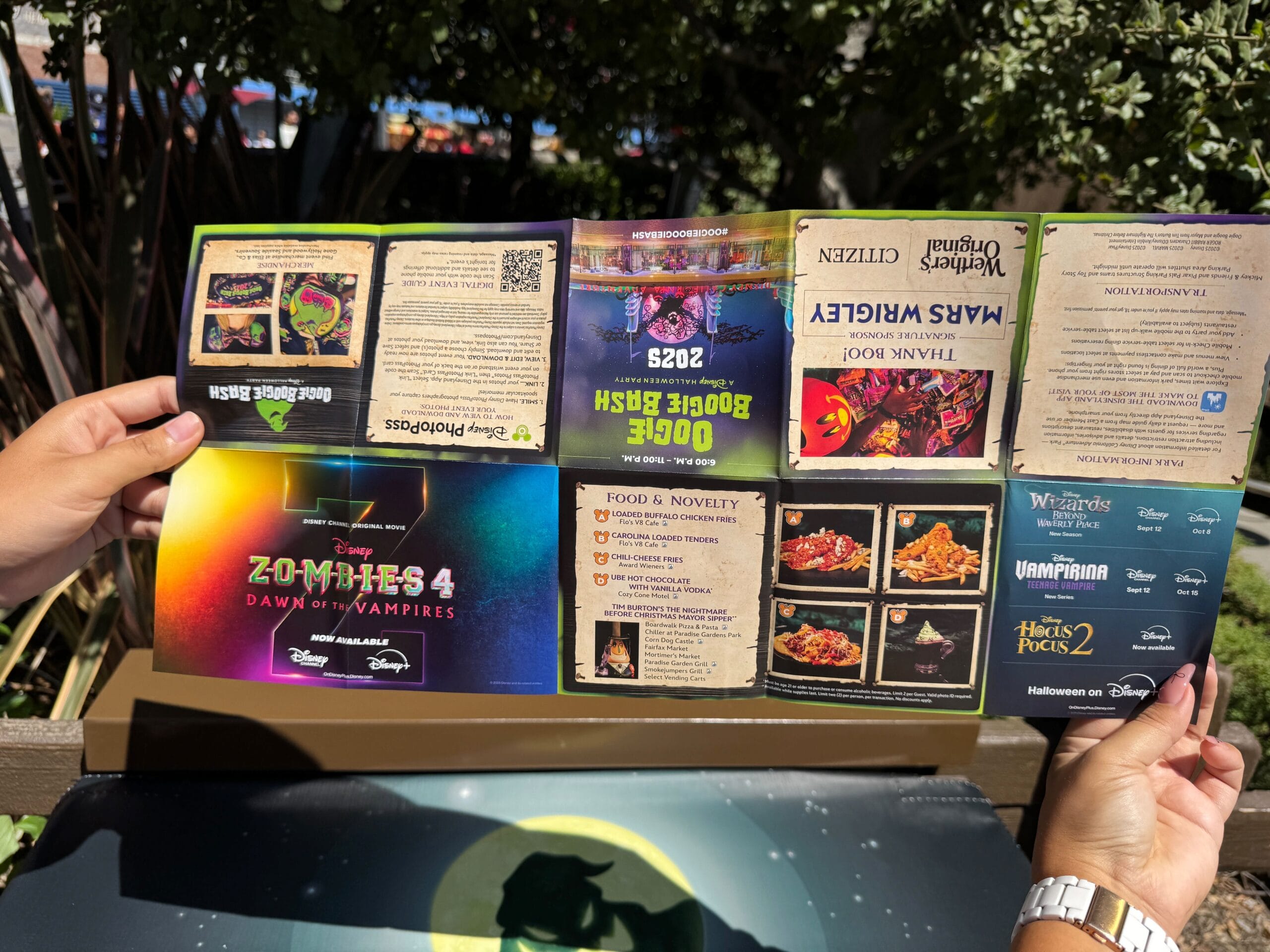 Person holding an outdoor event menu with upside down text and images; a wristband is visible in the sunlight’s shadows.
