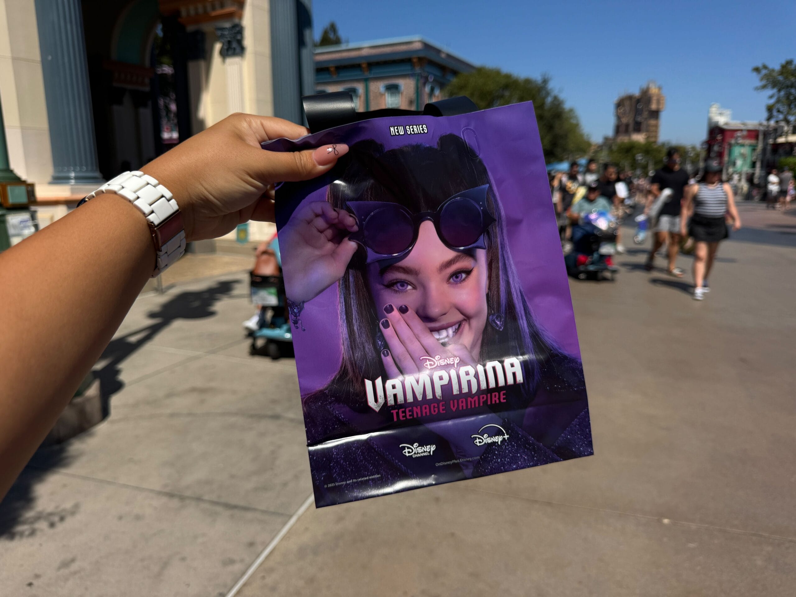 A hand holds a purple Disney "Vampirina: Teenage Vampire" promo bag and event map in a sunny theme park walkway.