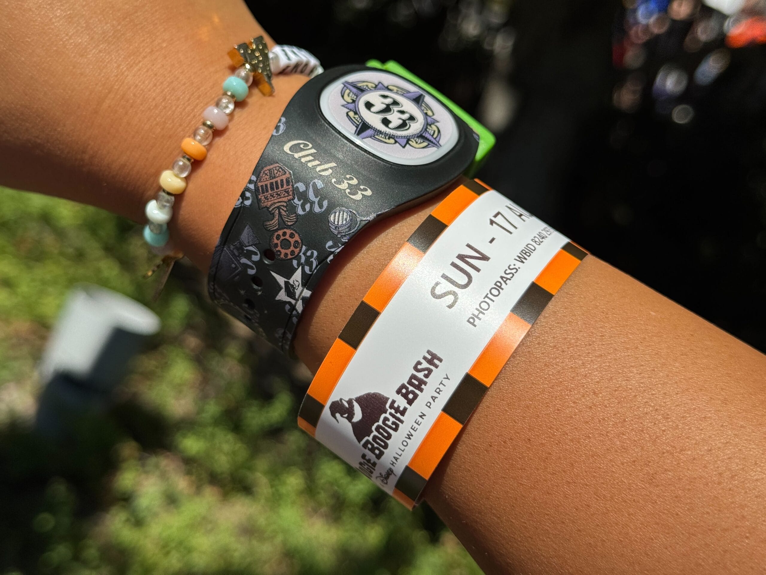 A wrist with a colorful beaded bracelet, MagicBand, and 2025 Oogie Boogie Bash event wristband gleams in sunlight.