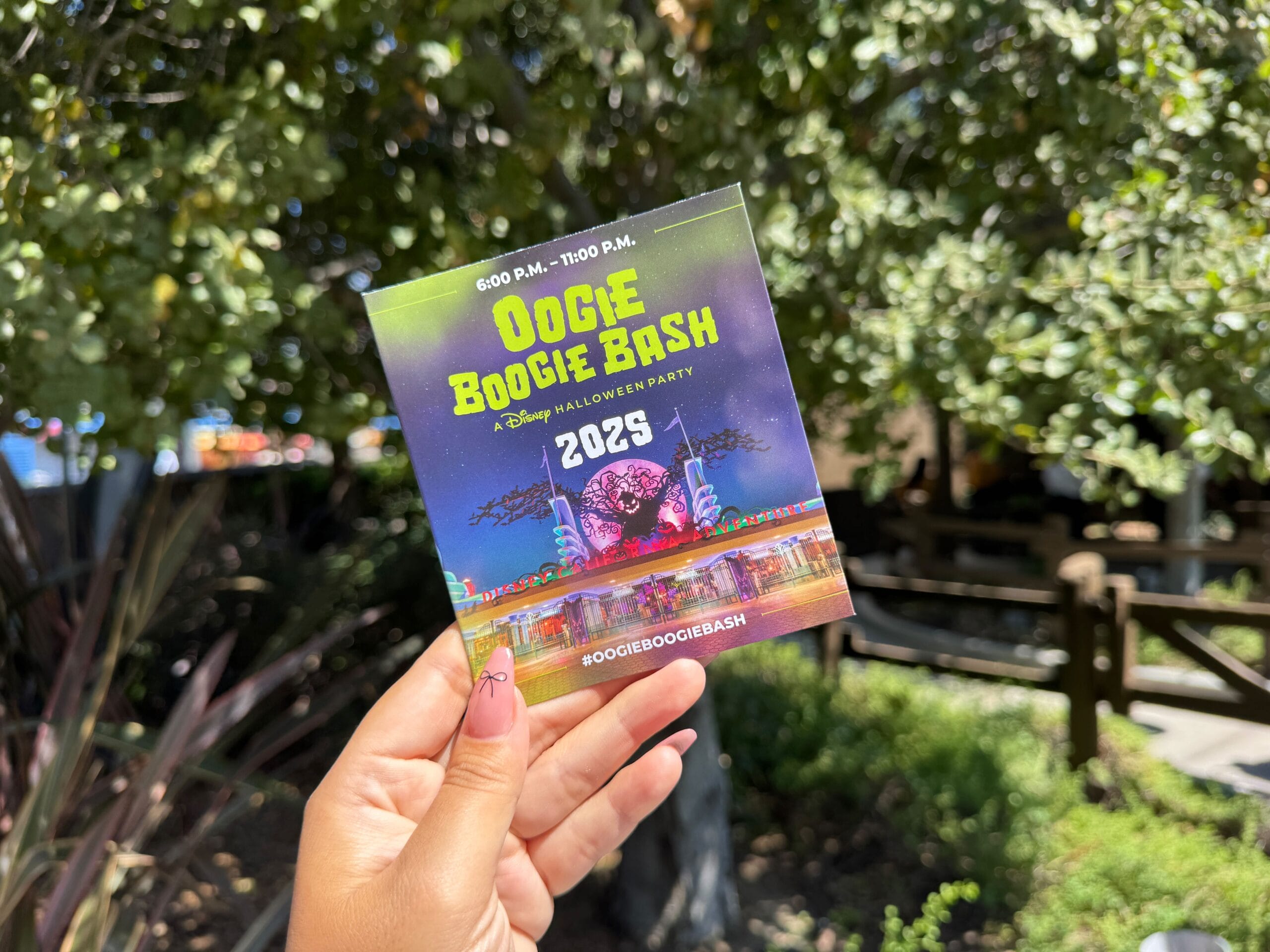 Hand holding 2023 Oogie Boogie Bash ticket with colorful design, trees, sunlight, and event map in the background.