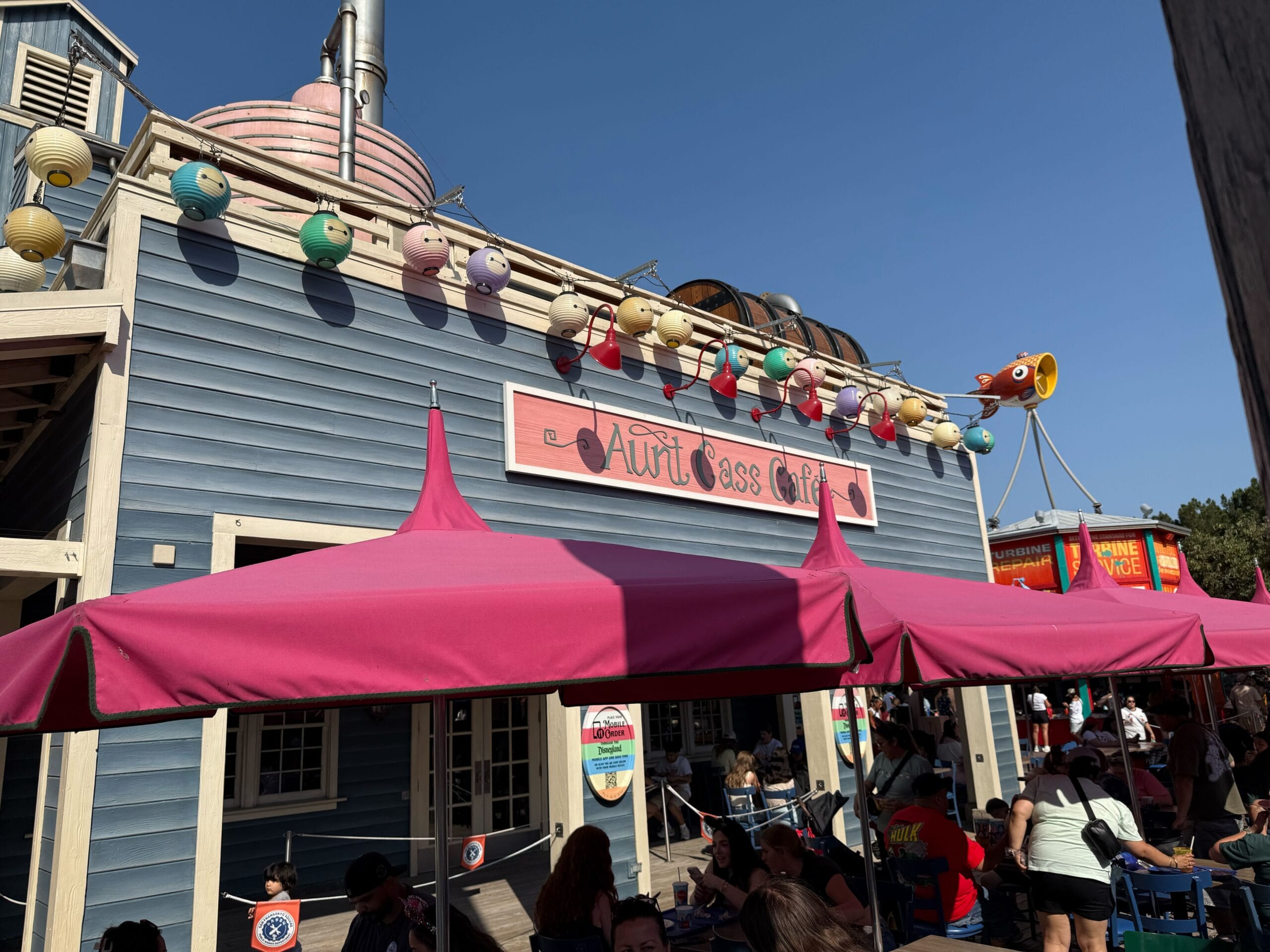 People gather outside "Aunt Cass Café" under pink umbrellas; colorful lanterns hang above the blue building.