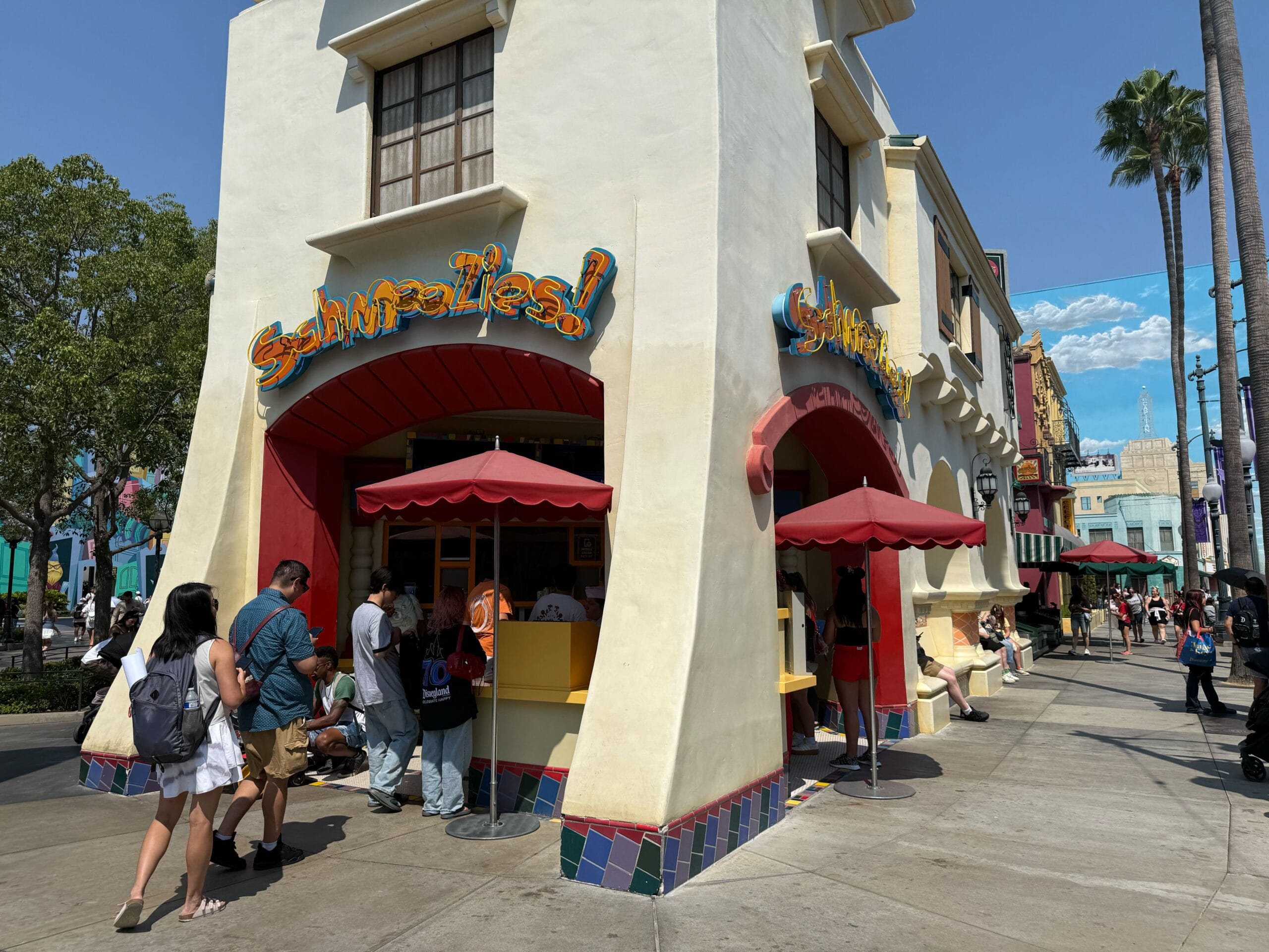 People stand outside a colorful corner snack stand with “Snacks!” signs, shaded by red umbrellas on a sunny day.