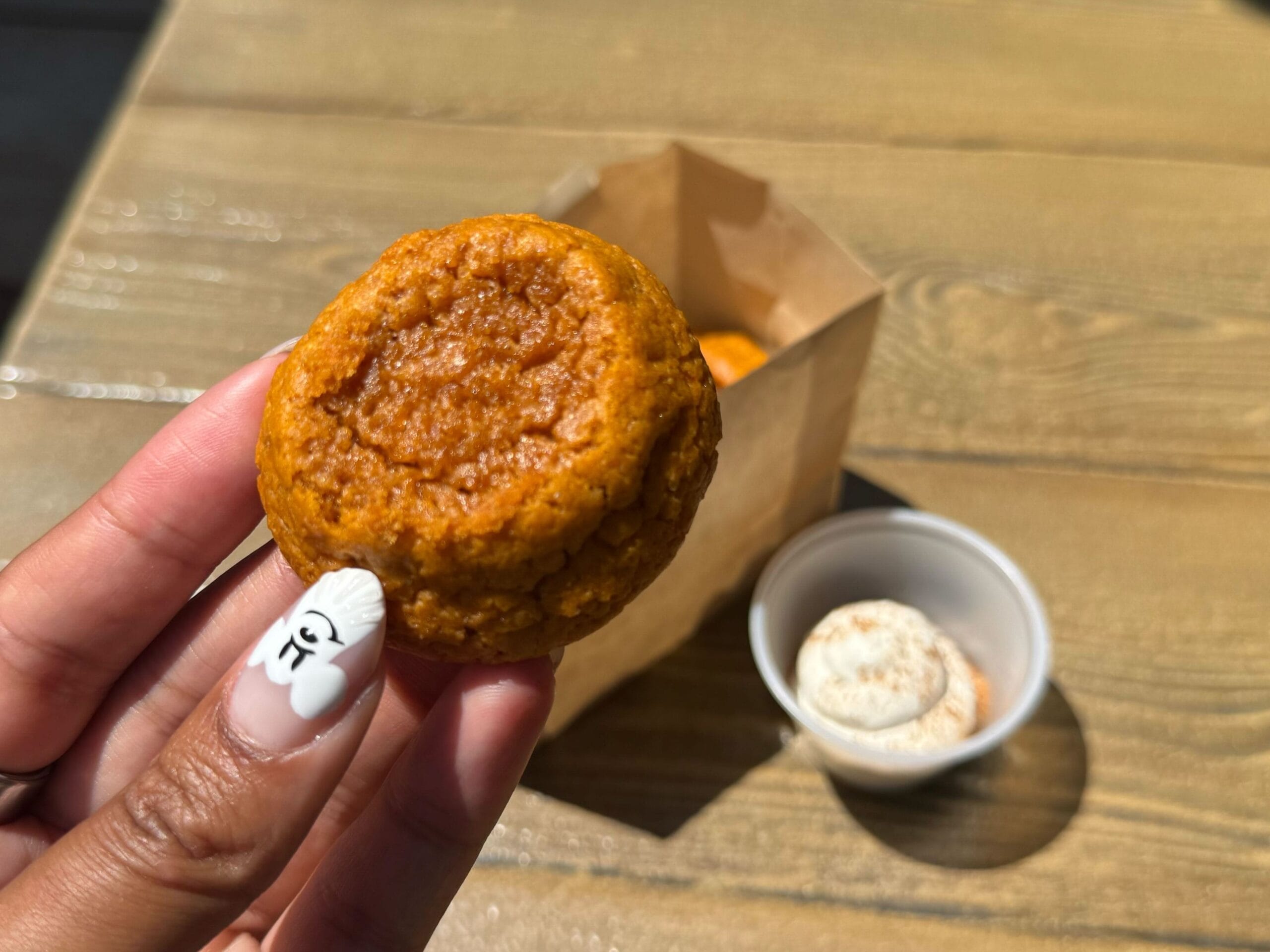 A hand holds a small orange muffin; Pumpkin Cookies and cream cheese dip sit on a wooden table in sunlight.