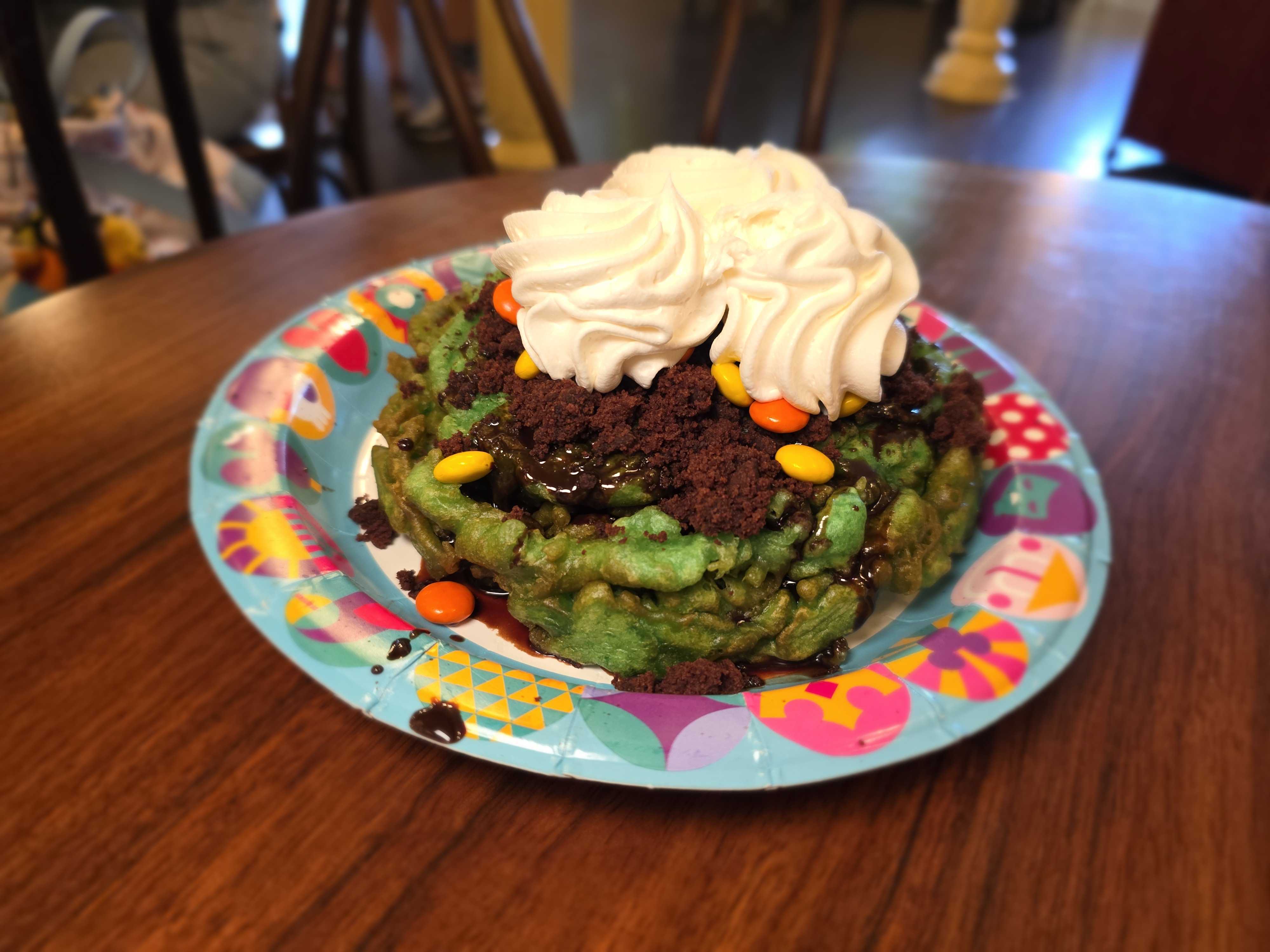A Disneyland Halloween Monster Funnel Cake: green waffle with whipped cream, chocolate syrup, cookies, and candy on a festive plate.