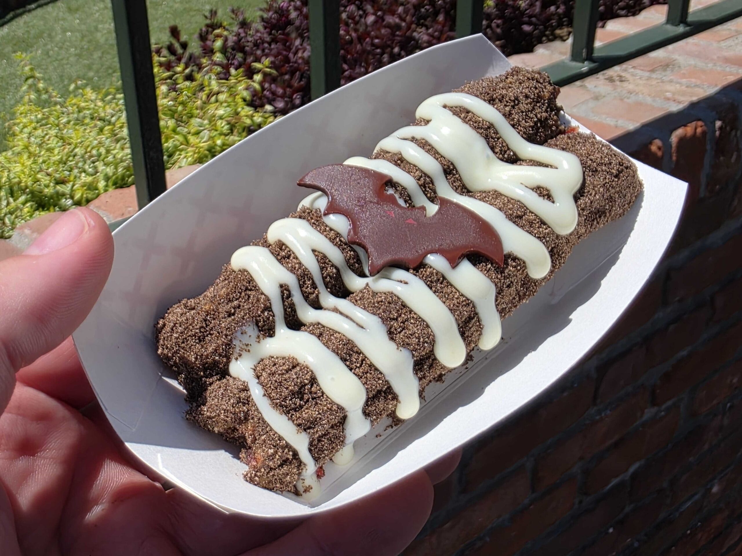 A hand holds Disneyland Halloween Churros with white icing, cookie crumbs, and a chocolate bat in a paper tray.