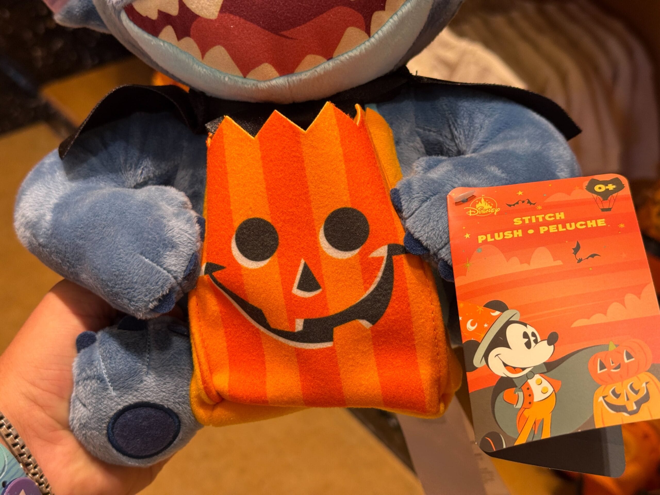 A plush Stitch toy holding a pumpkin pouch, featuring a Halloween Mickey Mouse tag—perfect Disneyland Halloween merchandise.