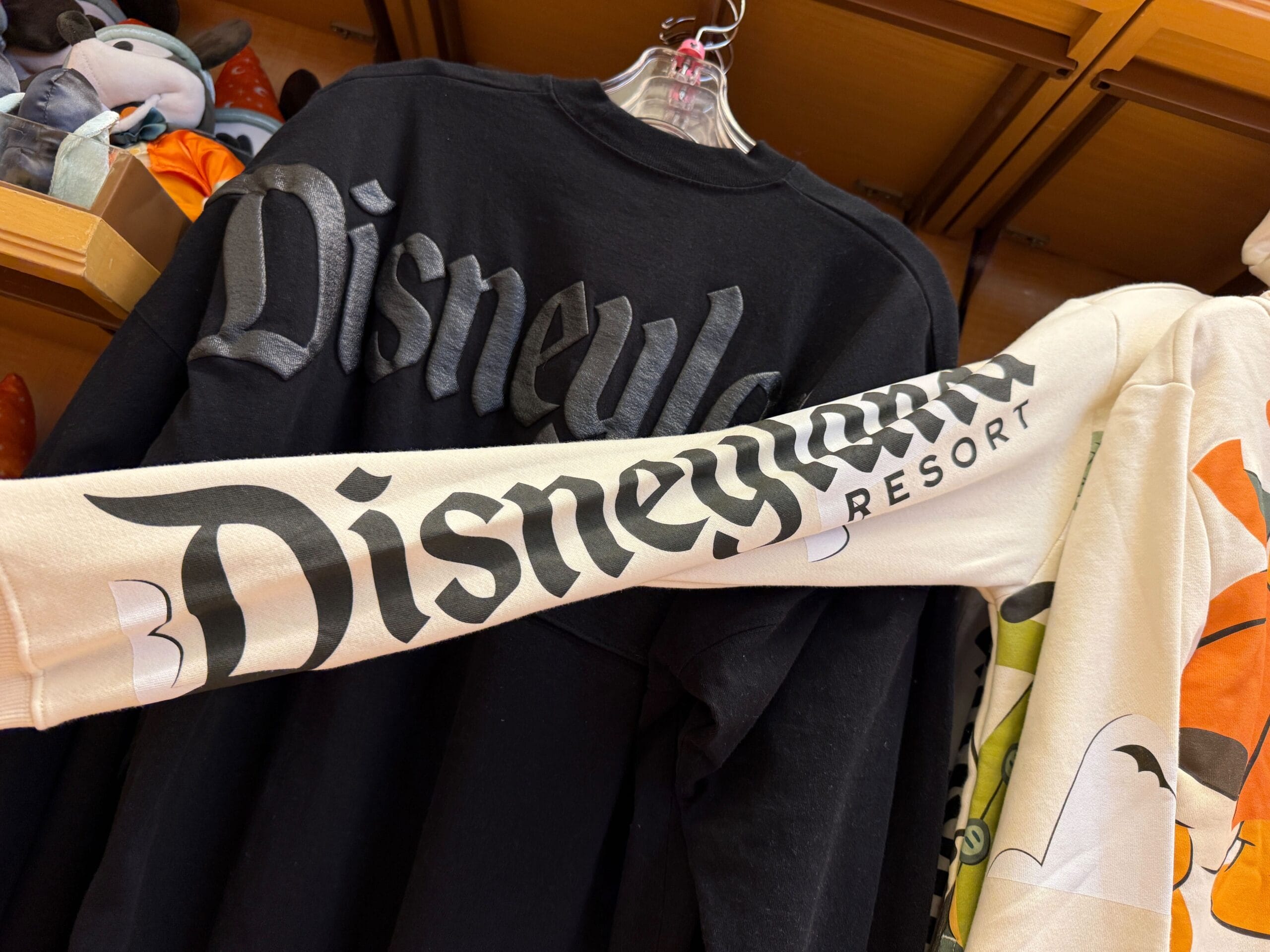 A black Disneyland Halloween merchandise sweatshirt hangs next to a cream Disneyland Resort shirt sleeve in a store.