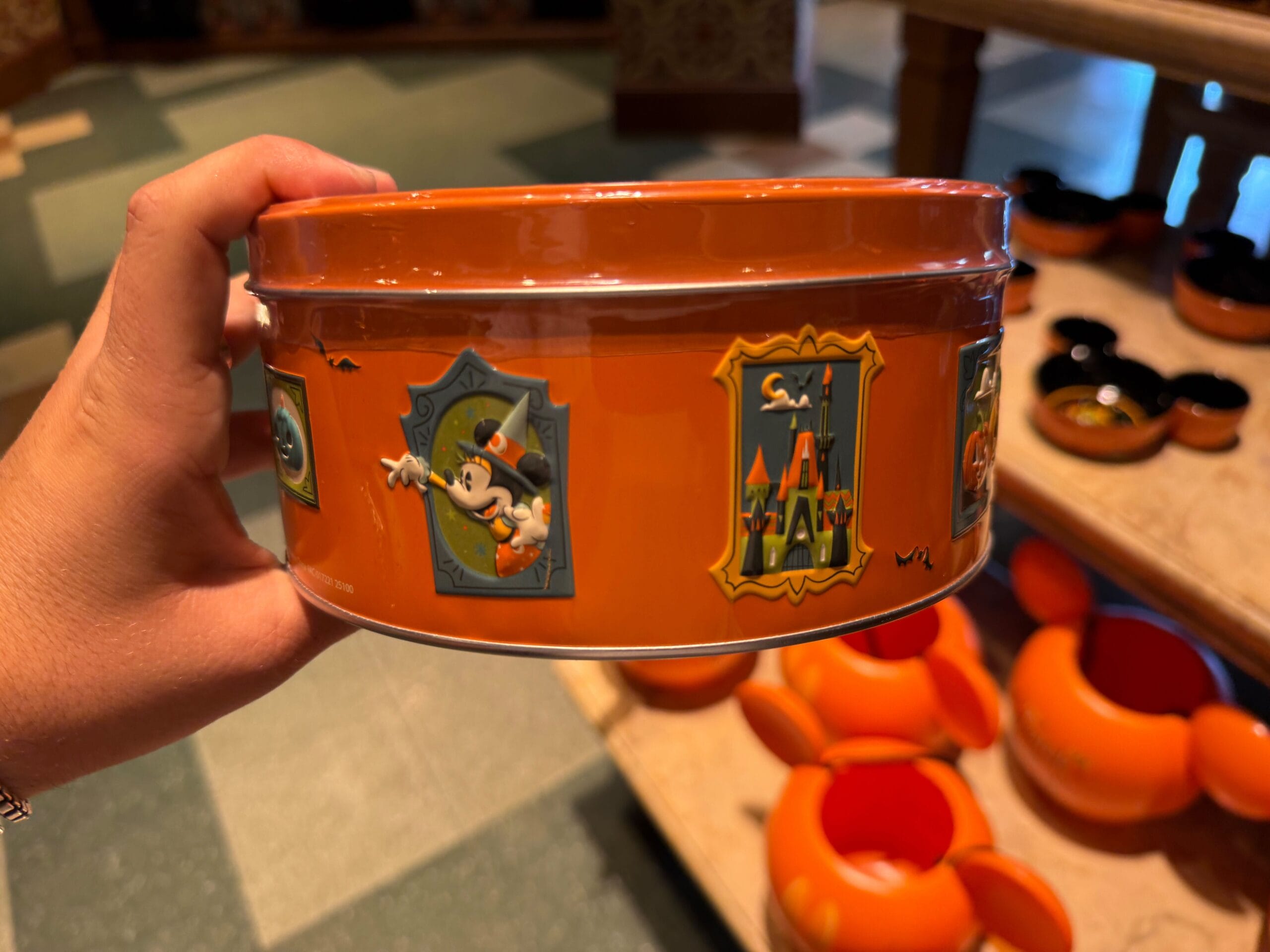 A hand holds an orange tin of Disneyland Halloween merchandise, decorated with festive Disney designs in a store.