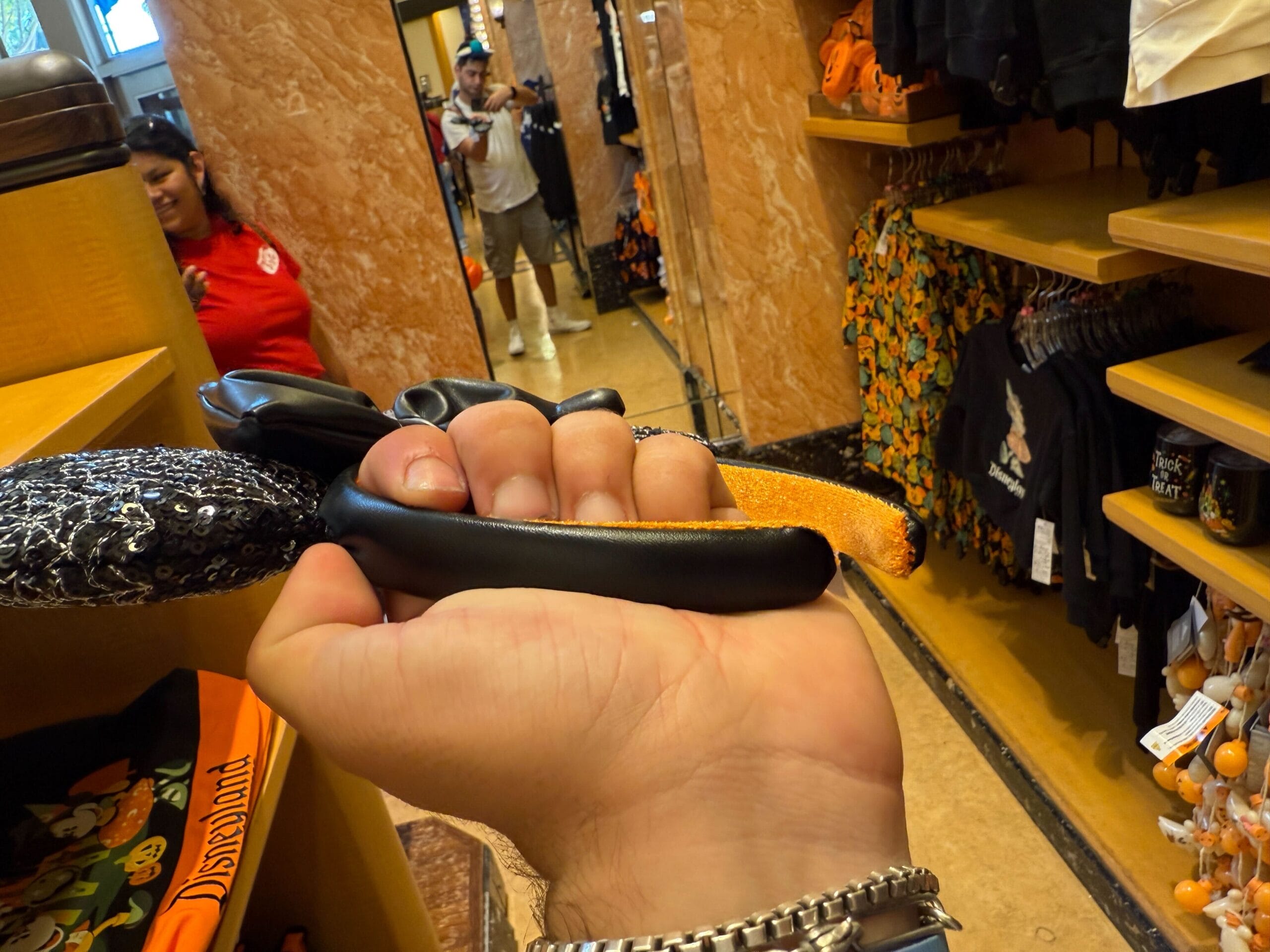 A hand holds Mickey Mouse ears headband in a store filled with Disneyland Halloween merchandise on the shelves.