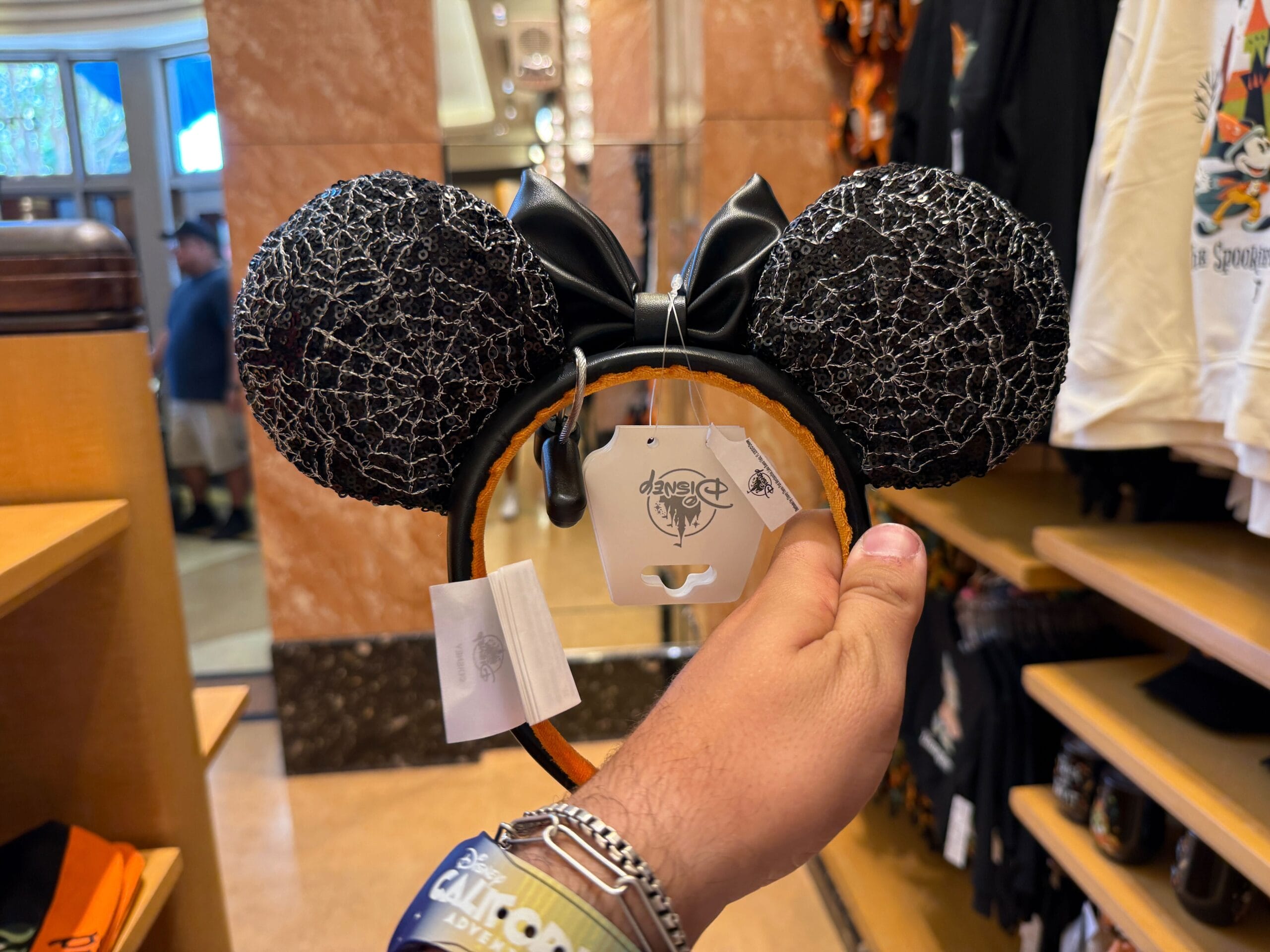A hand holds black Minnie Mouse ears with a spiderweb bow amid Disneyland Halloween Merchandise in a festive store.