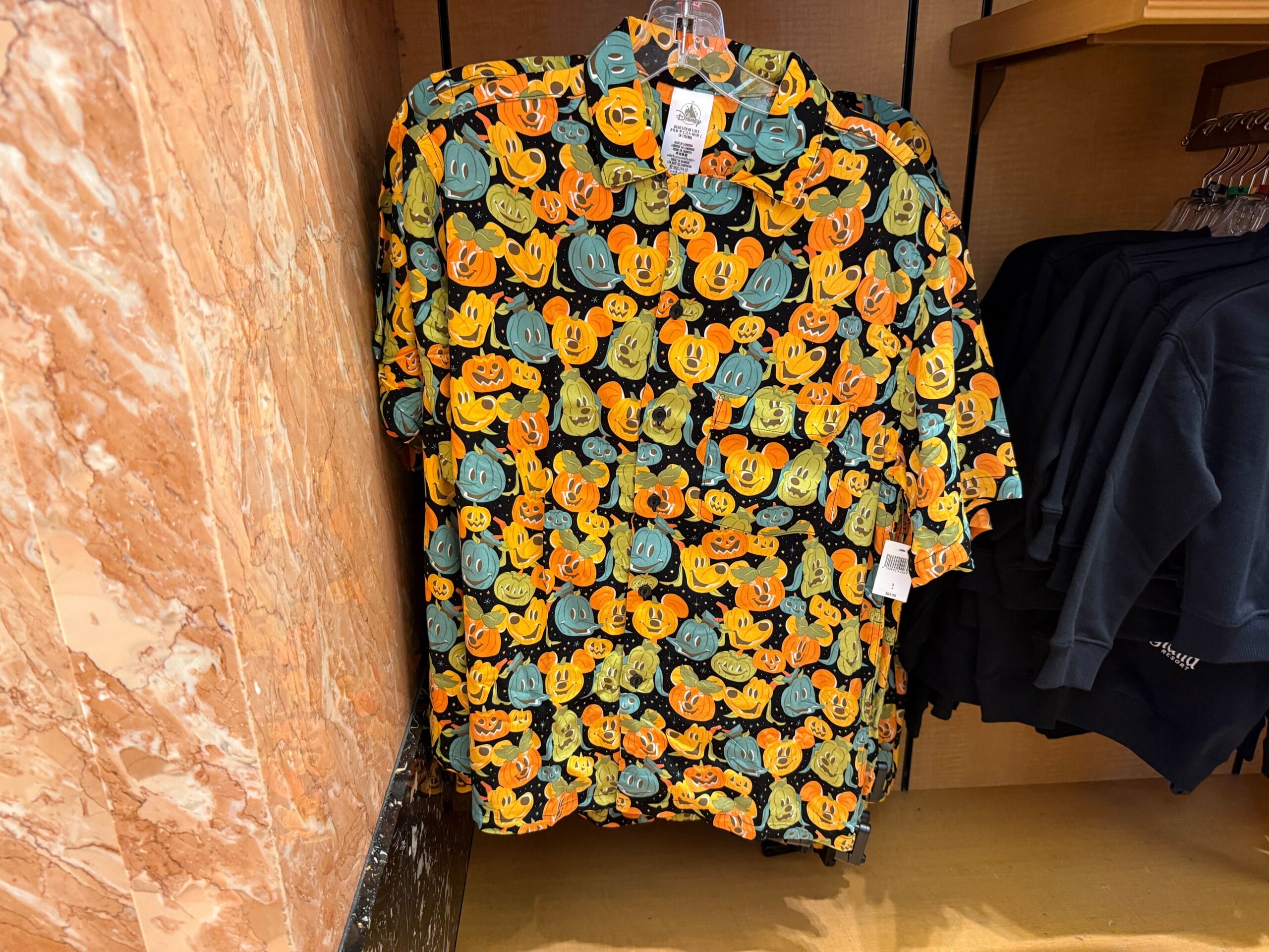 A button-up shirt with a colorful pumpkin Mickey Mouse pattern hangs on display among Disneyland Halloween merchandise.