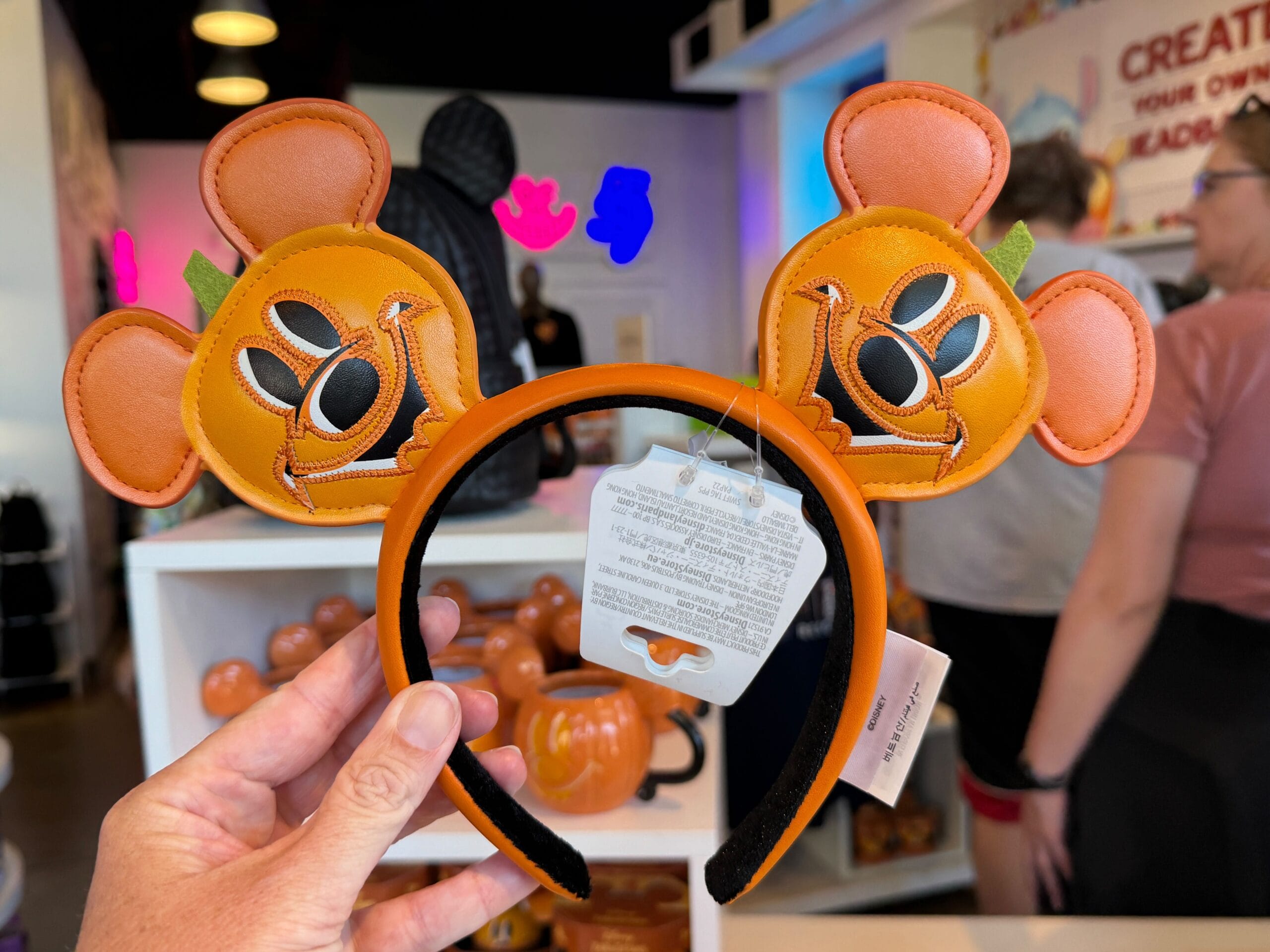 Orange Minnie Mouse ear headband with pumpkin design, a festive Pumpkin Merch find, held in-store among shoppers and pumpkin decor.