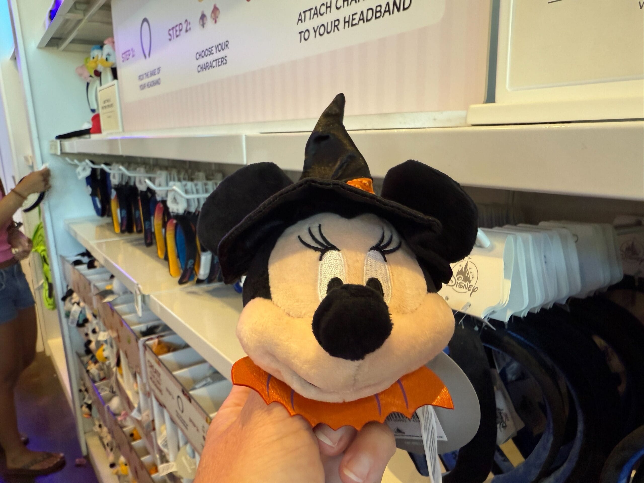 A hand holds a Minnie Mouse witch plush in a store aisle with Pumpkin Merch and headbands on display.