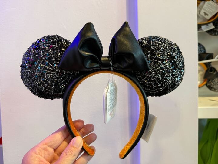 A hand holds a Minnie Mouse ear headband with black spiderweb ears, a black bow, and festive Pumpkin Merch flair.