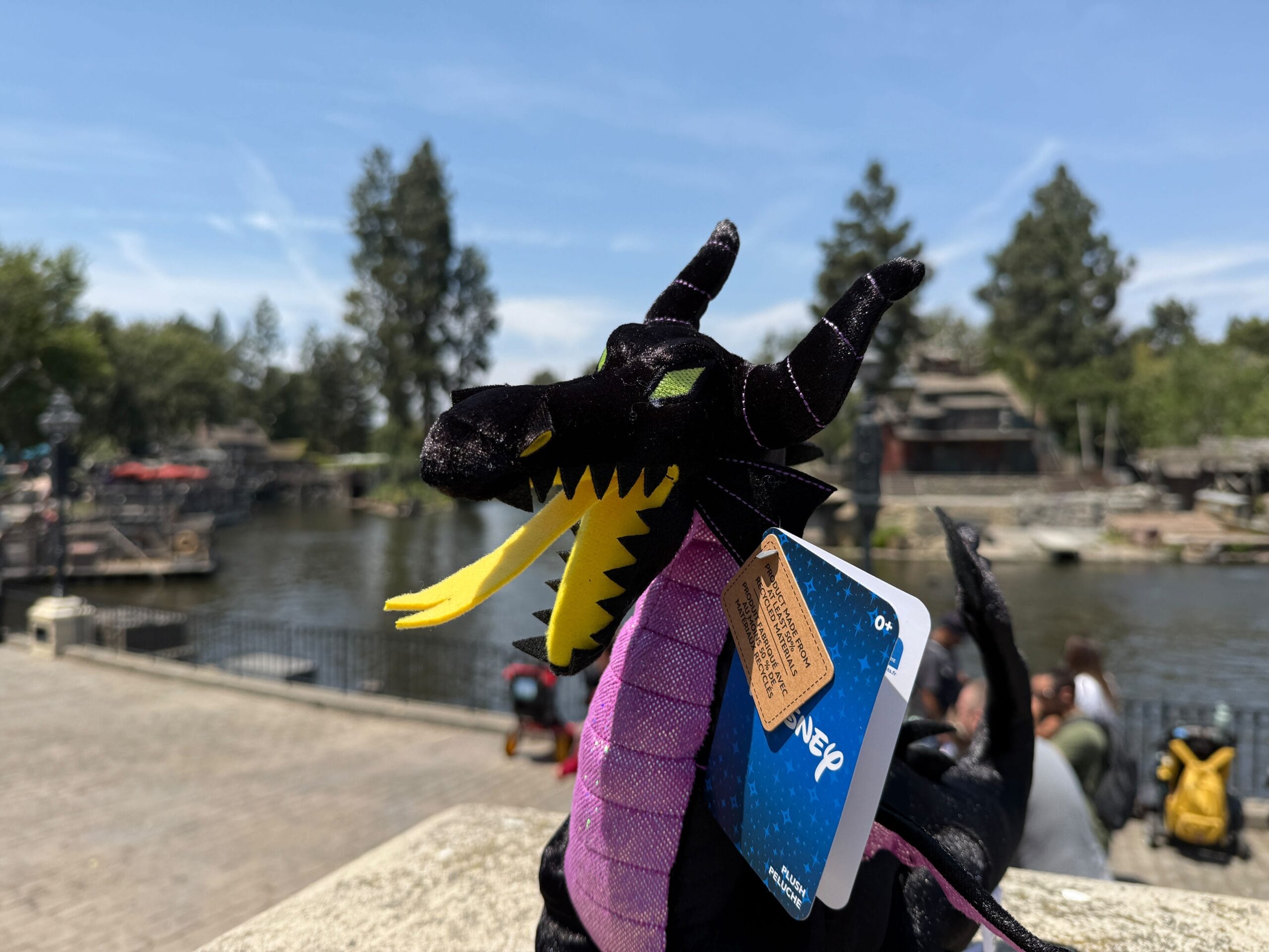 A Maleficent Dragon Plush with tags sits outdoors near a river on a sunny day at Disneyland Resort.