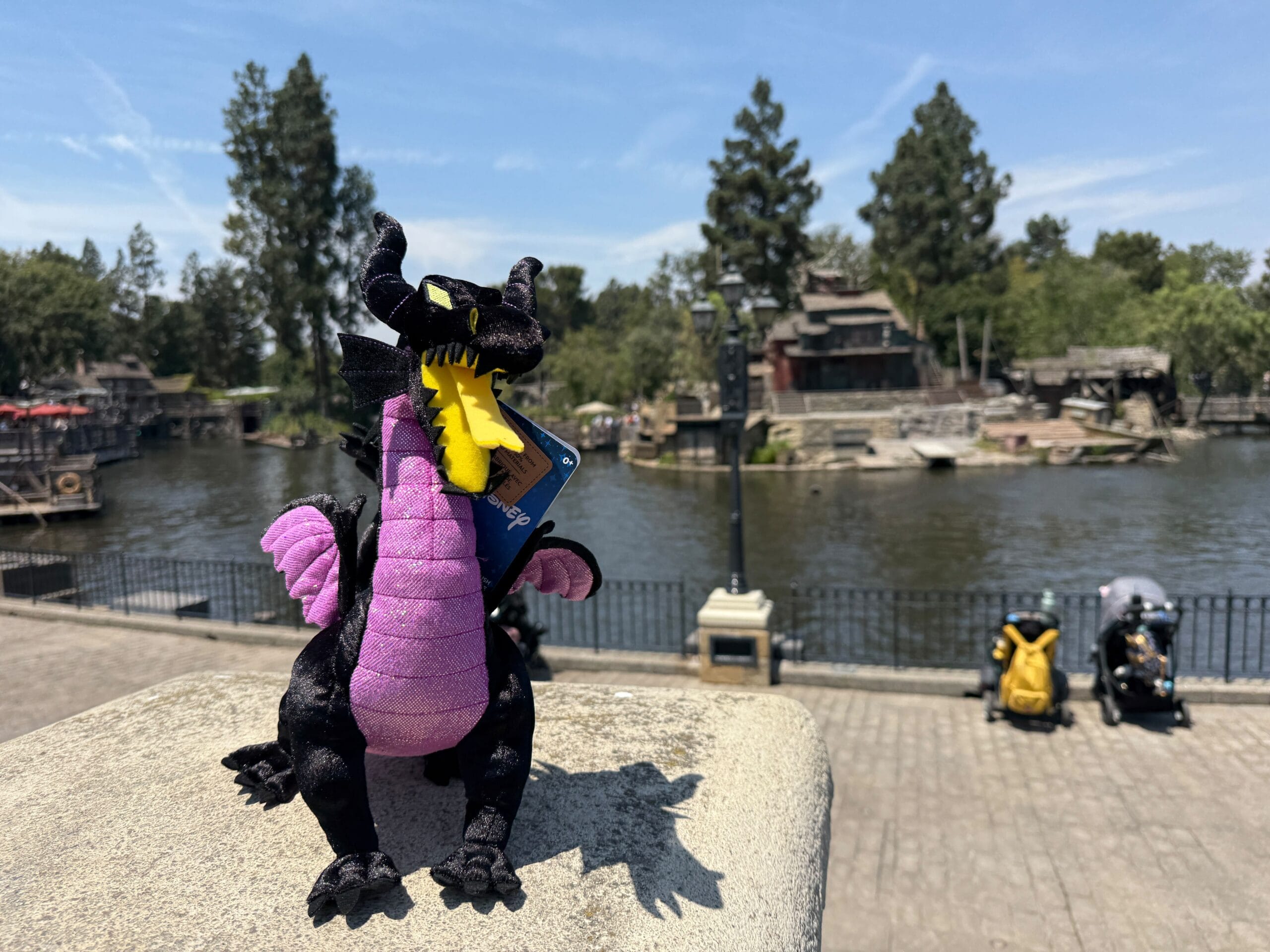 A Maleficent Dragon Plush holding a card stands on a stone ledge by the riverside at a park, trees and strollers behind.