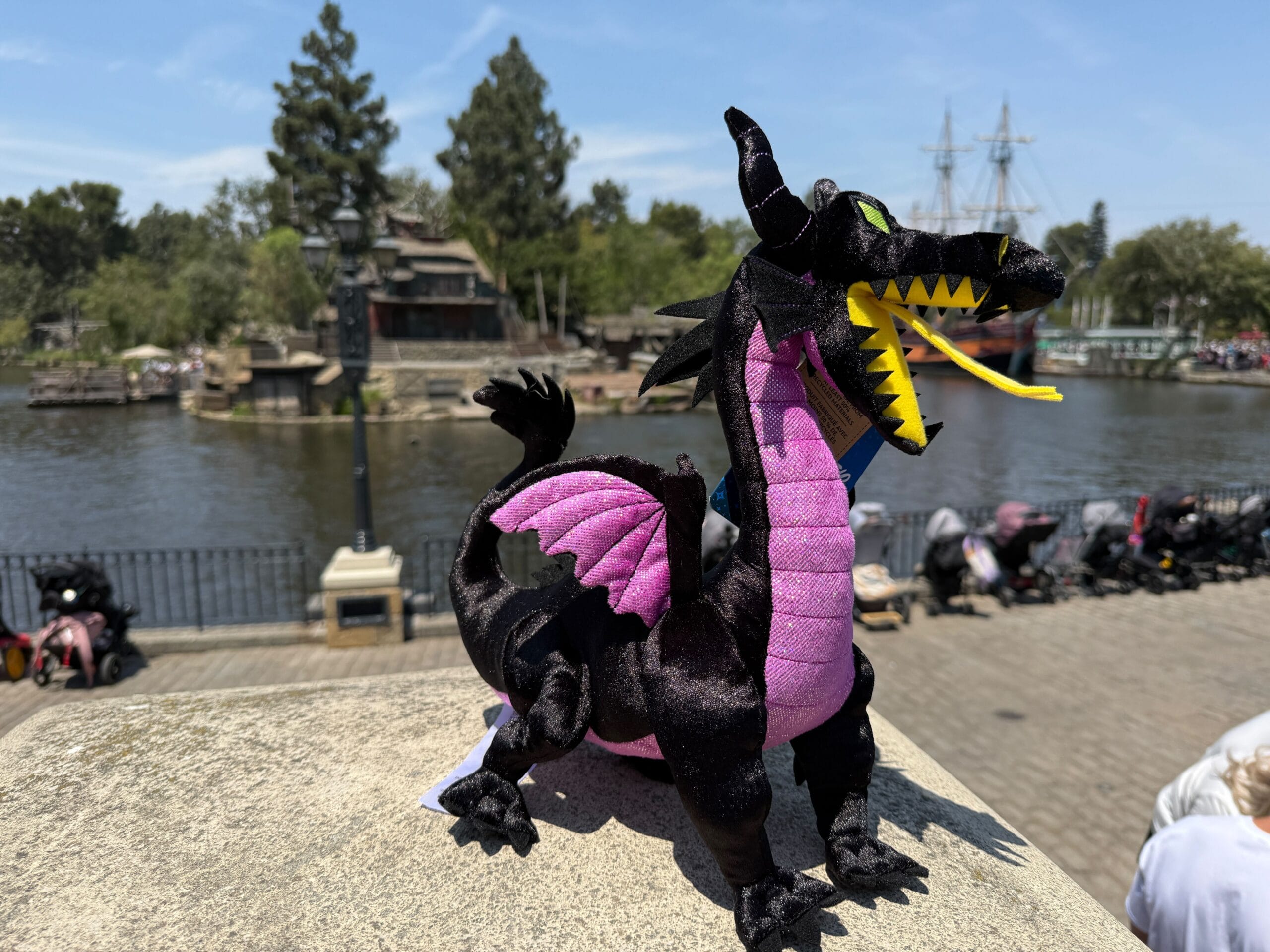 A Maleficent Dragon Plush sits on a stone ledge by a riverside, with trees and a ship at Disneyland Resort in the background.