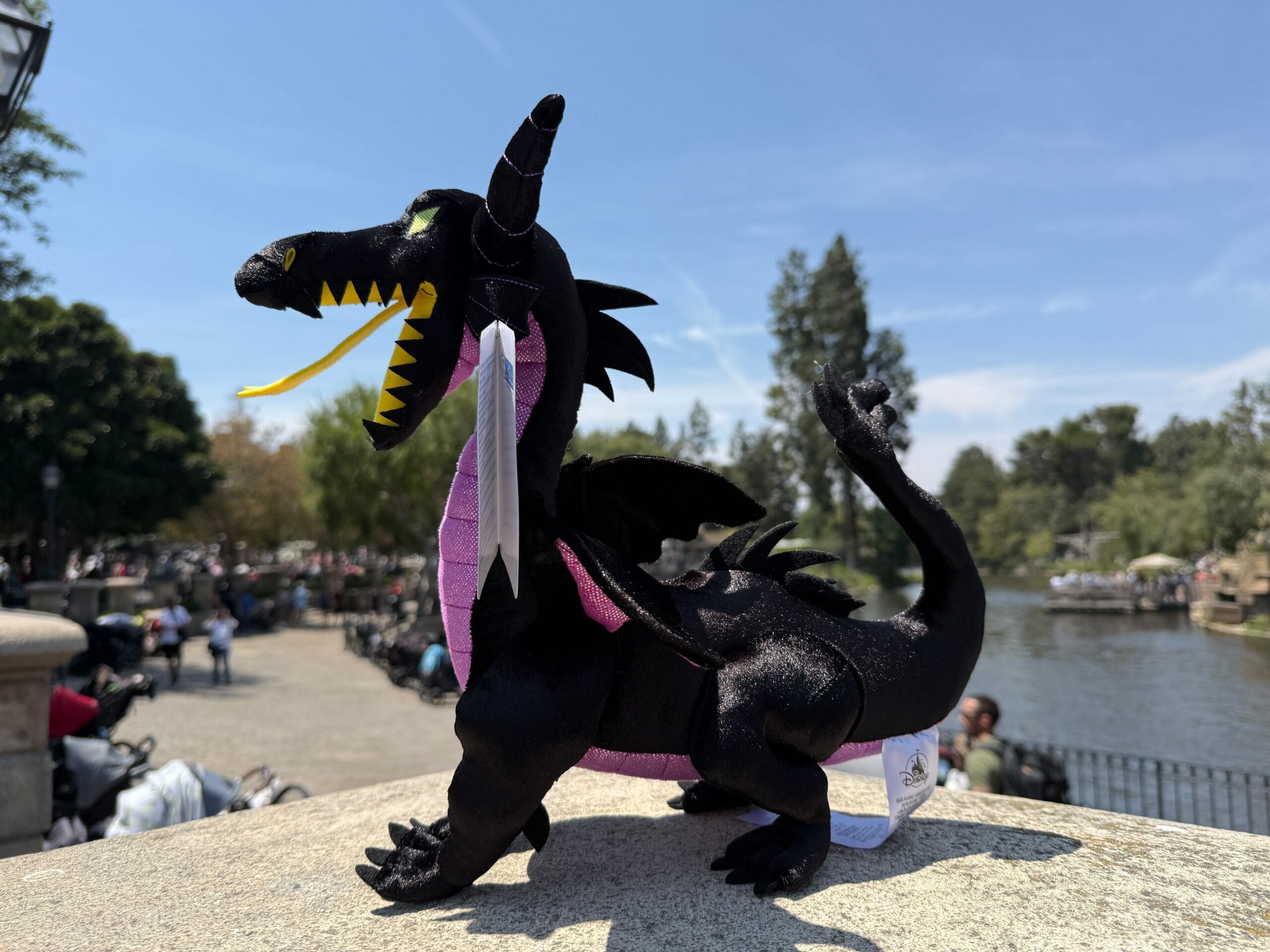 A Maleficent Dragon Plush sits on a stone ledge outdoors near water and trees under a blue sky.