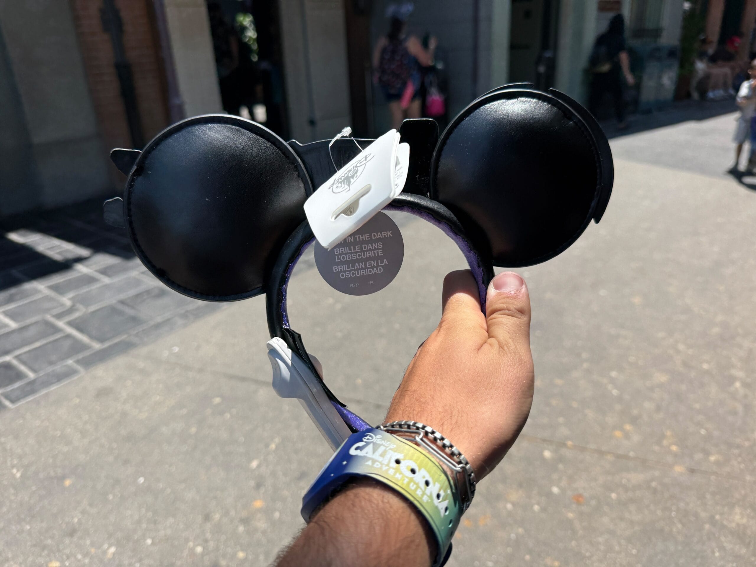 A hand holds a Fantasmic Ear Headband with a tag, outside on a sunny day at Disneyland Resort.